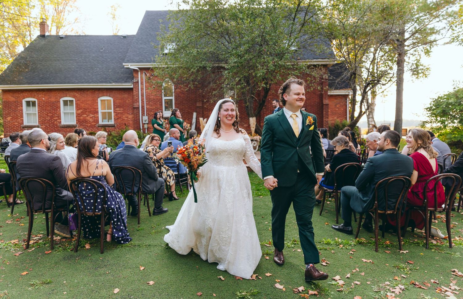 Wedding guests gather on a lawn outside a red brick building during an outdoor ceremony.