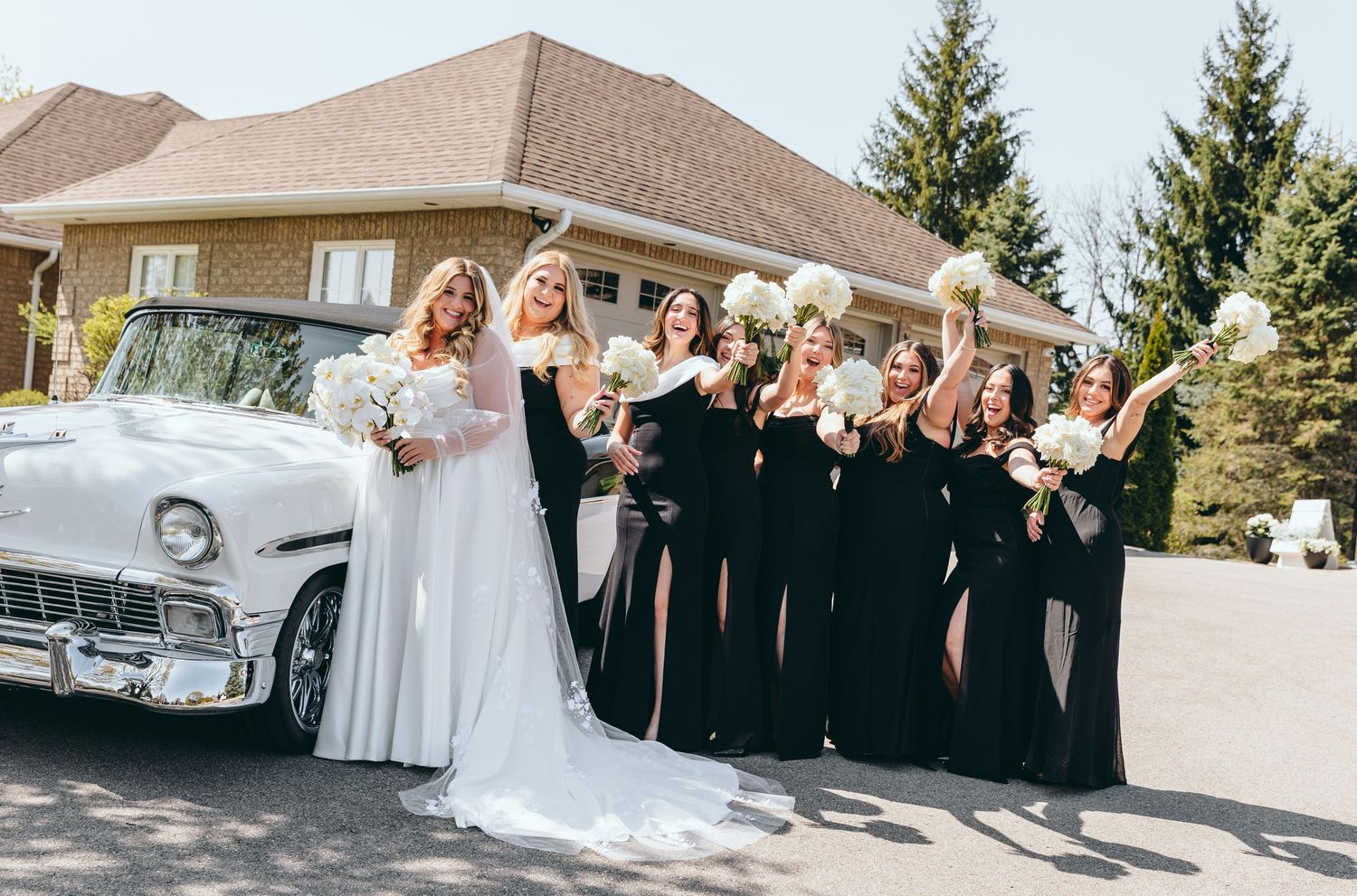 Wedding party poses joyfully next to a classic white vintage car.