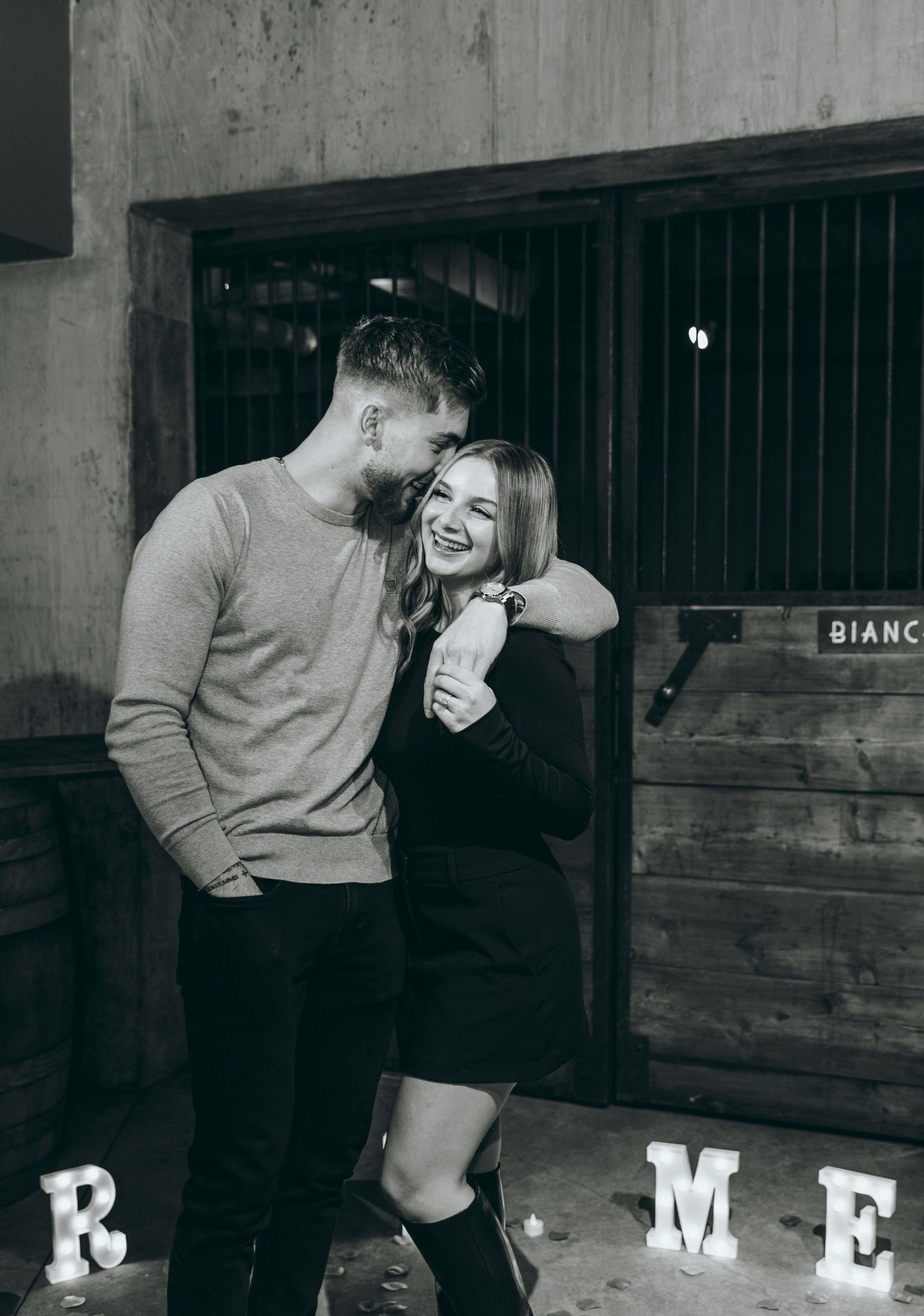 Romantic moment captured in industrial space with 'MARRY ME' letters visible.