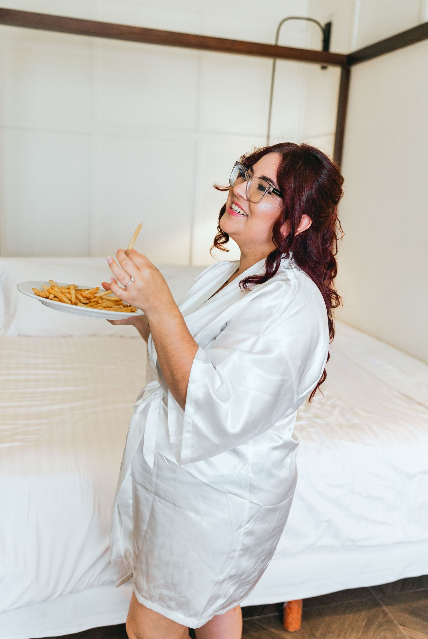 Person in white silk robe enjoys french fries while sitting on bed during wedding preparations.