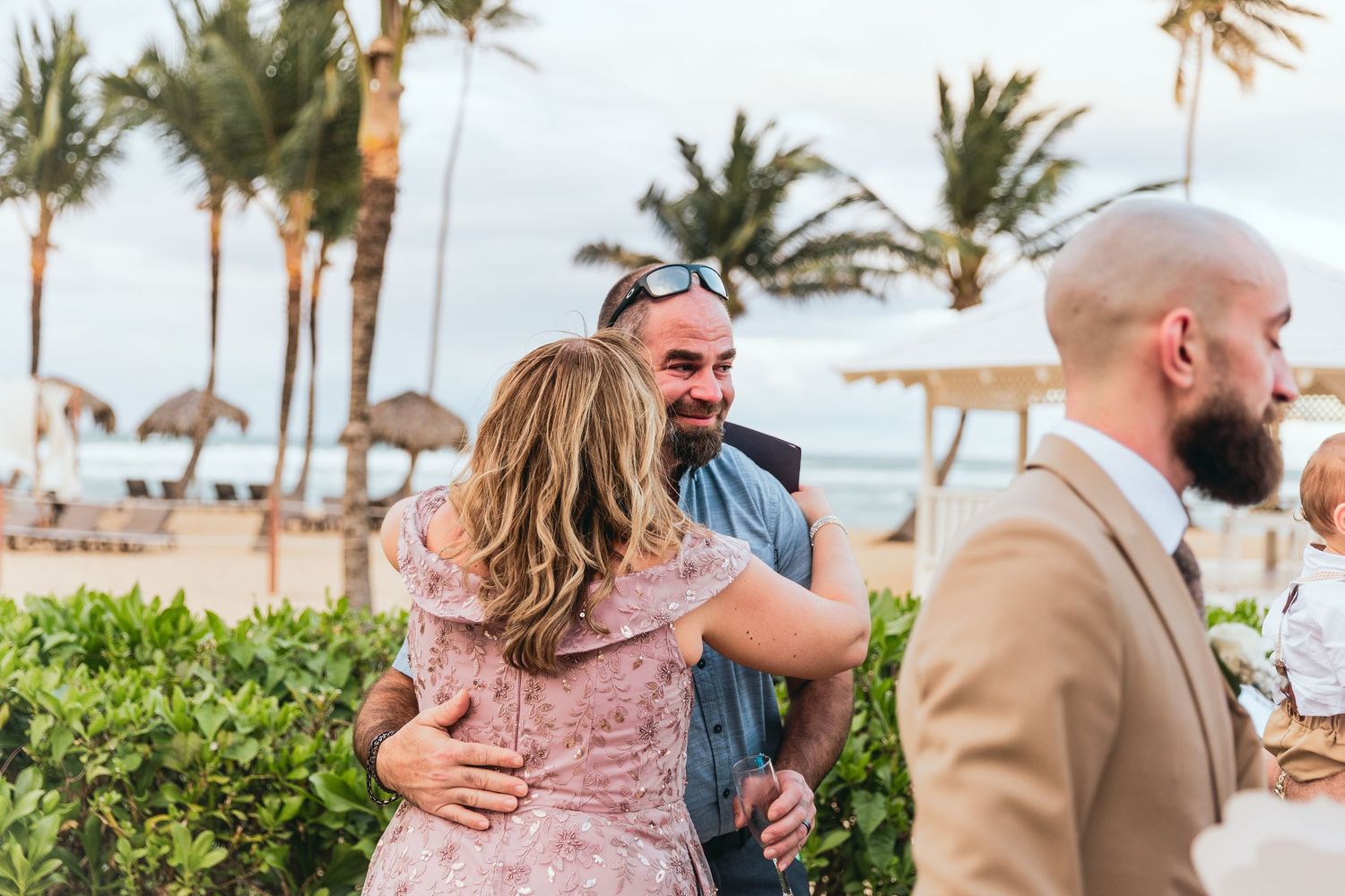 Guests embrace and celebrate during a beachside wedding reception with palm trees in view.