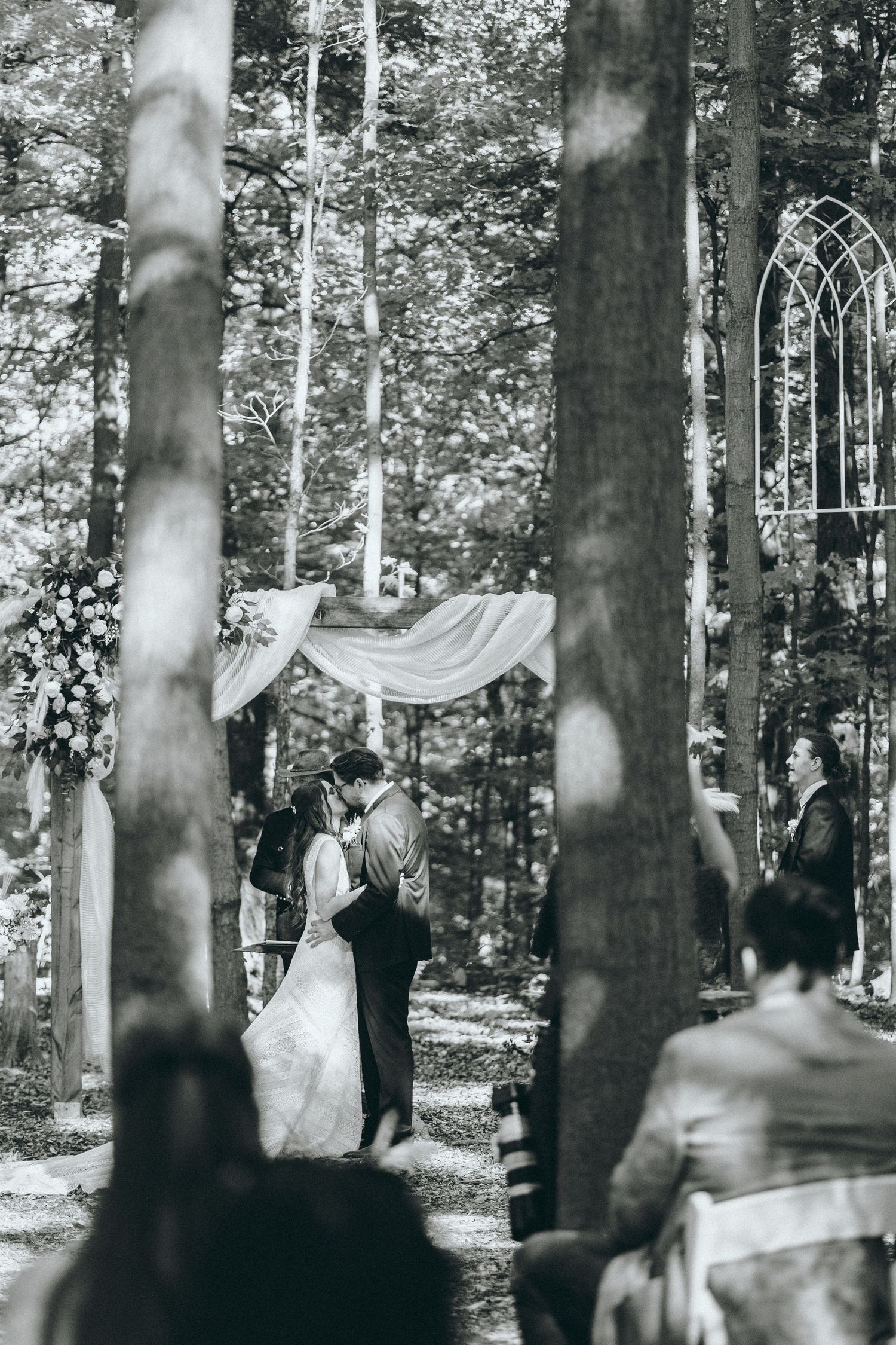 Black and white photo of an intimate forest wedding ceremony beneath flowing white fabric and tall trees.