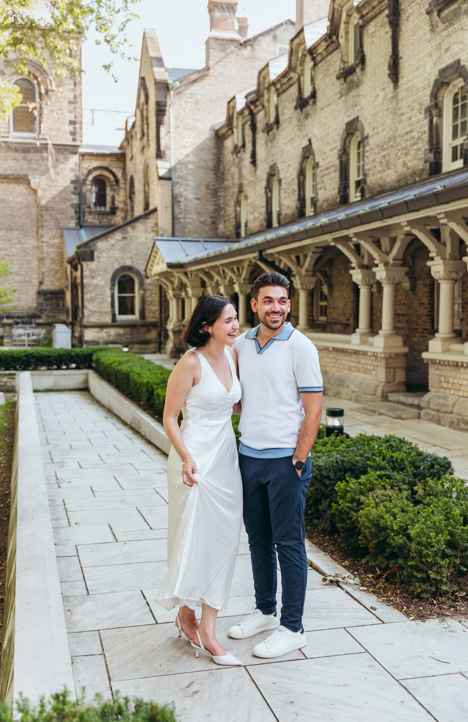 A couple shares a joyful moment along a stone colonnade walkway of a Gothic-style building.