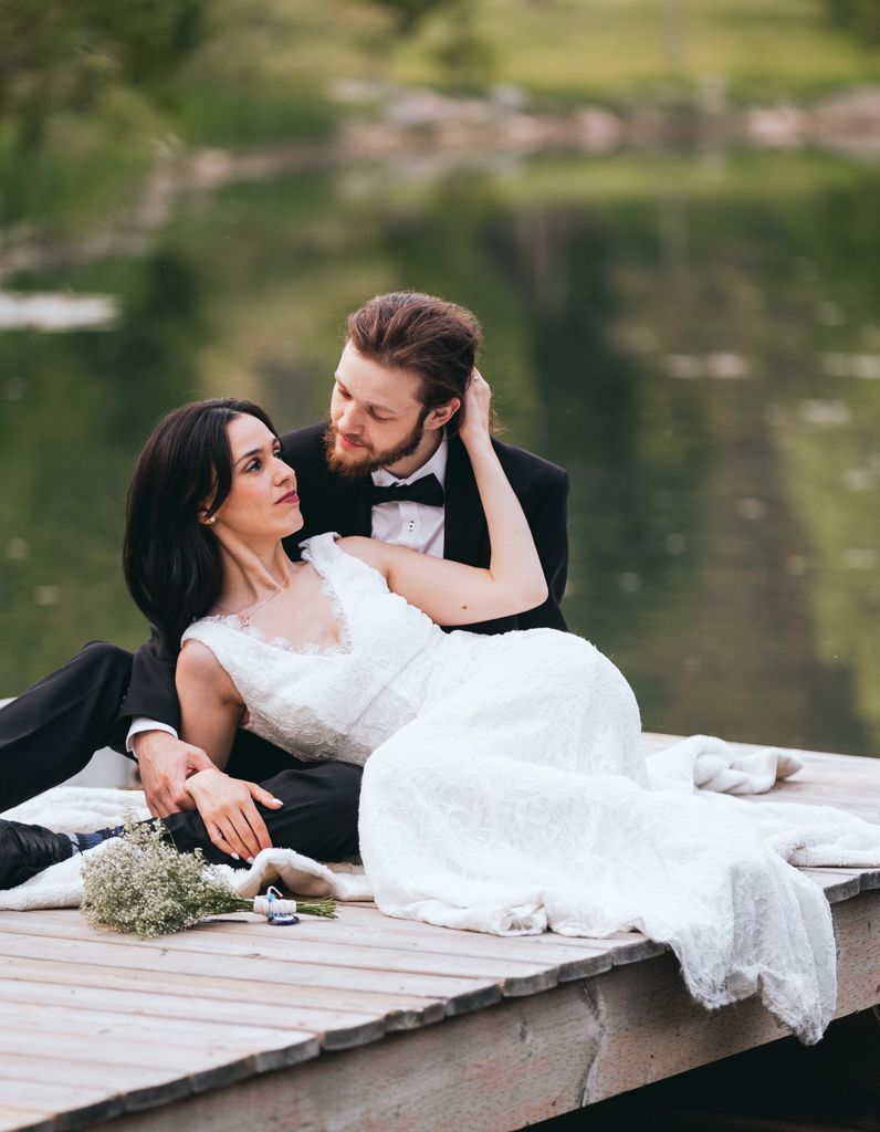 A couple in wedding attire shares an intimate moment while relaxing on a lakeside dock.