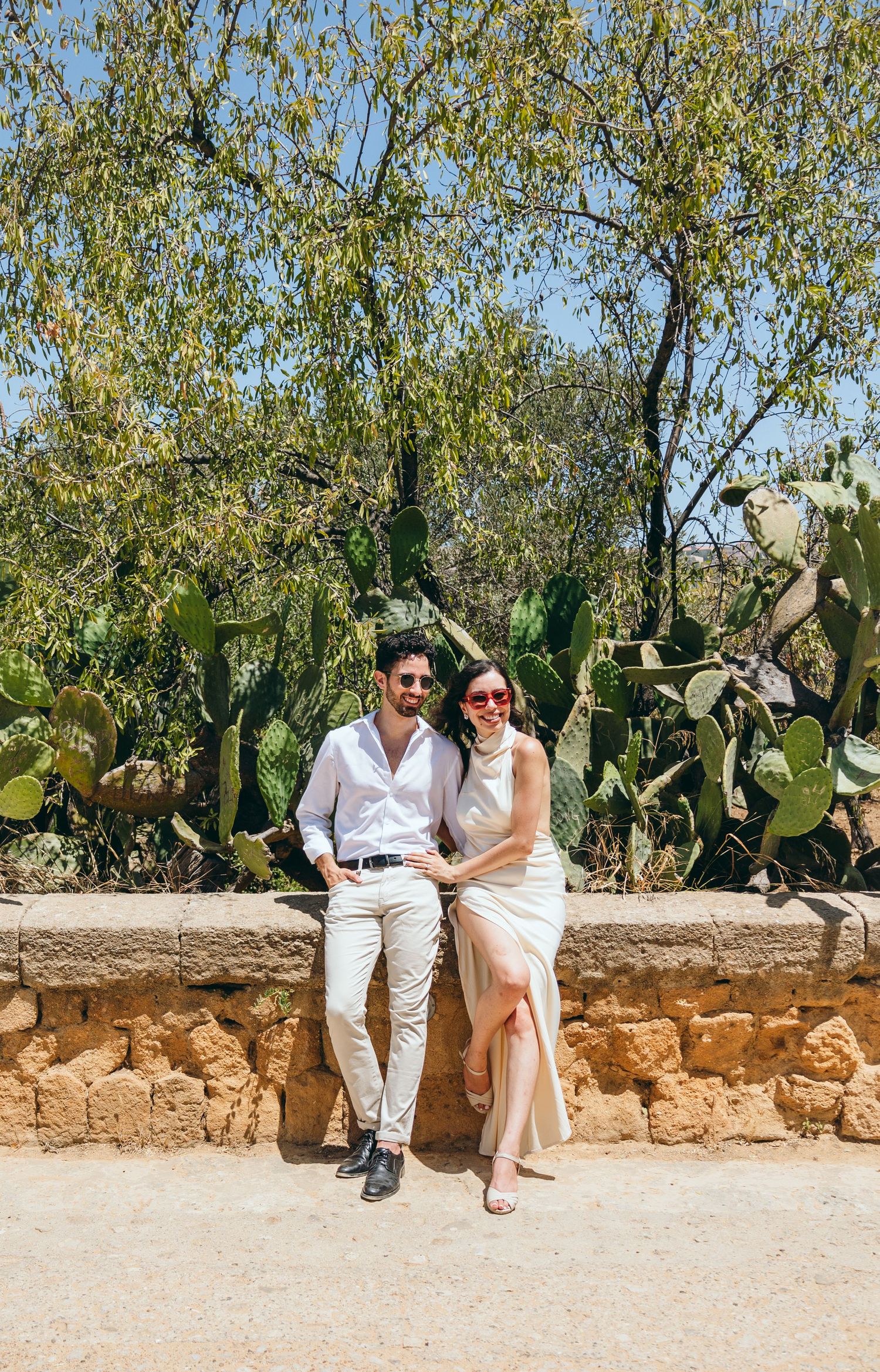 Stylish couple in white clothing sitting on rustic stone wall with cacti and olive trees.