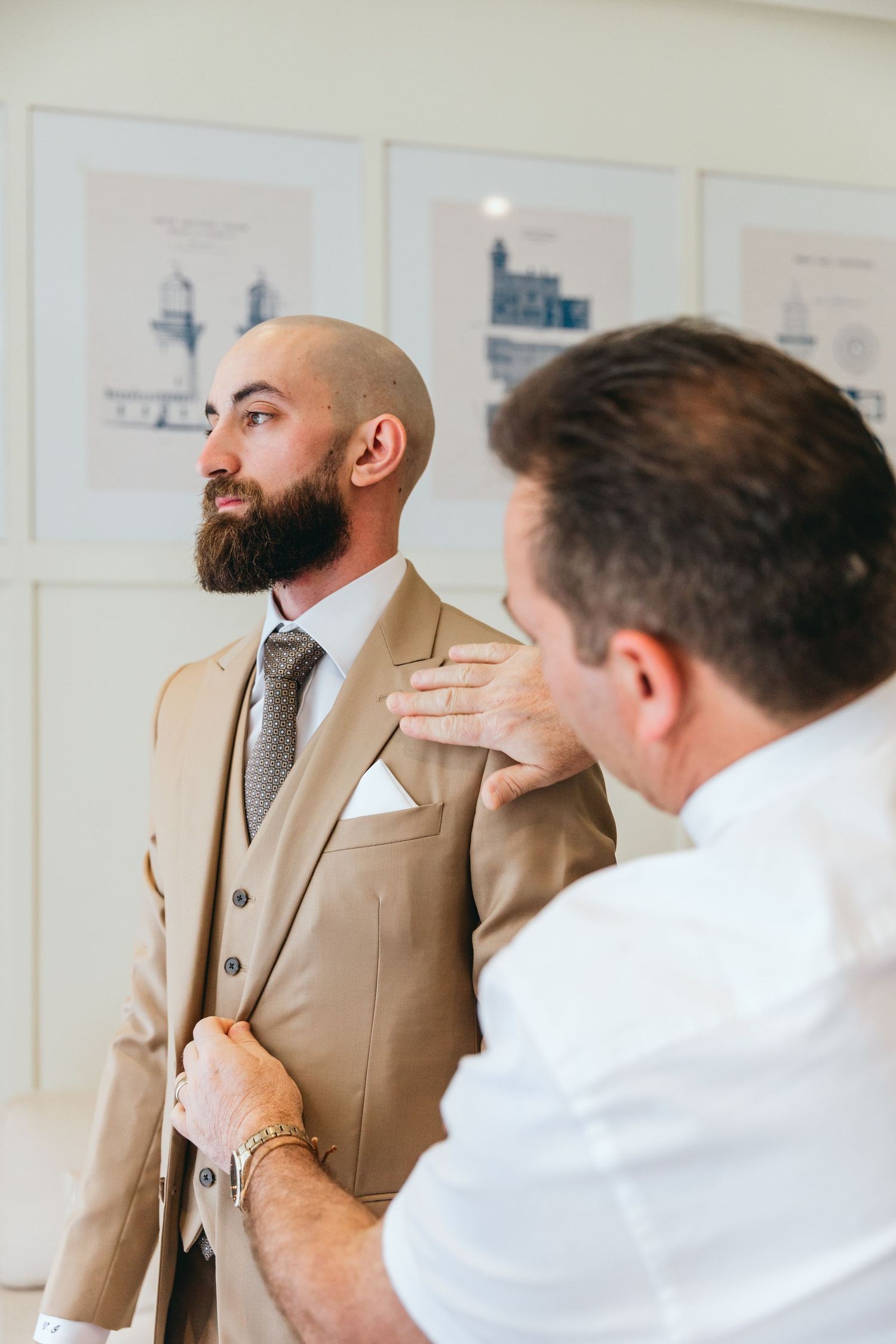 A groom in a tan suit gets final adjustments before the ceremony against a wall with framed artwork.