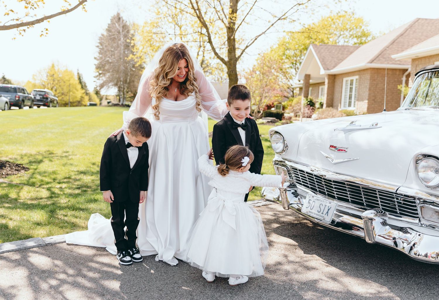 A bride in white dress stands with young attendants next to a classic car.