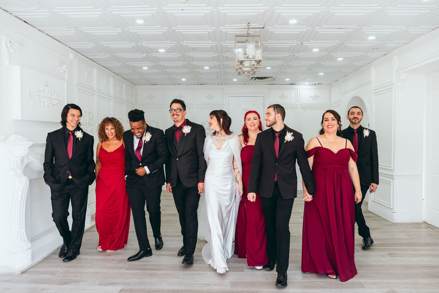 Wedding party walking and laughing together in a white room wearing burgundy and black formal attire.