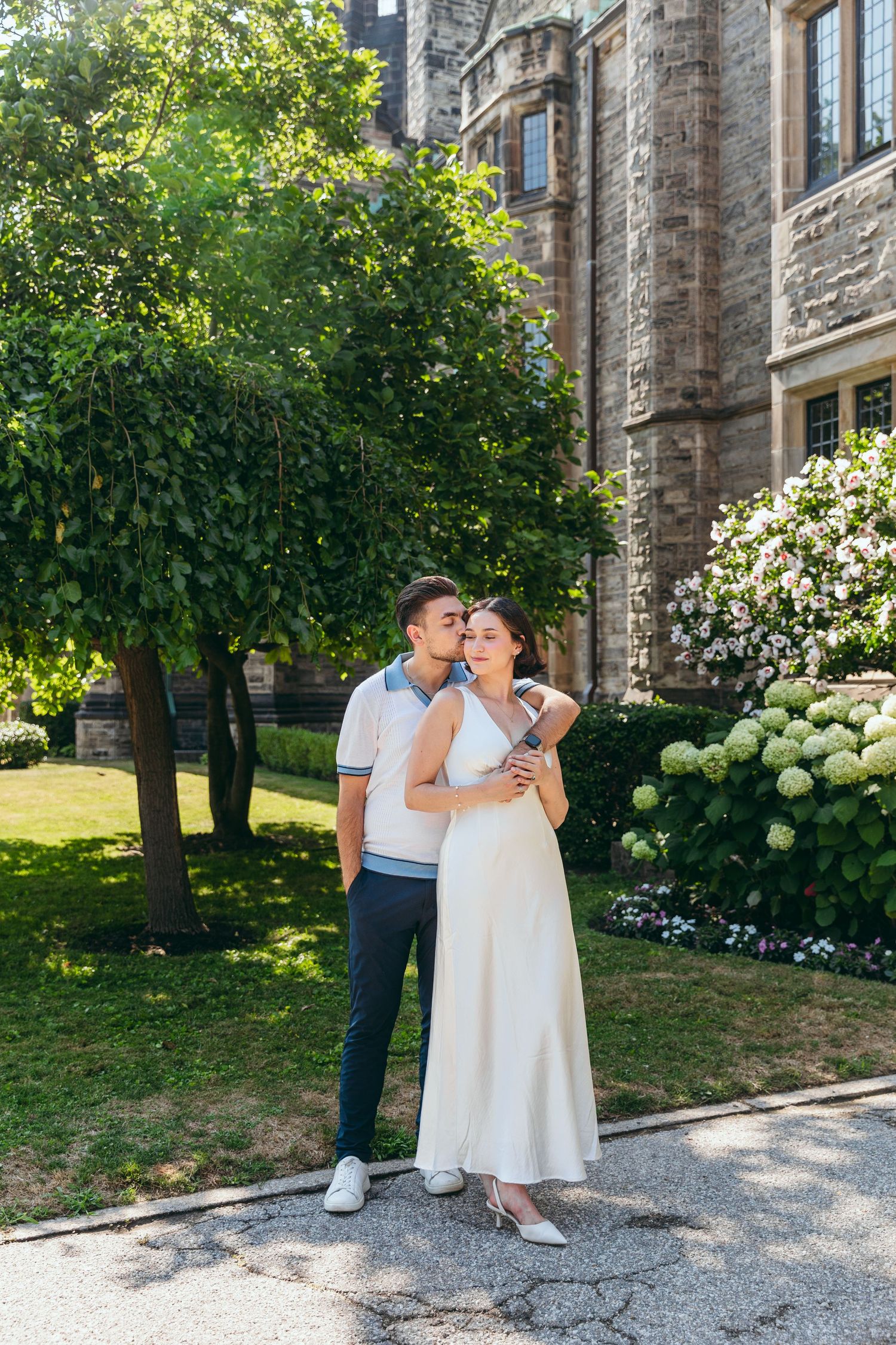 Couple embraces in garden setting with blooming hydrangeas and gothic architecture.