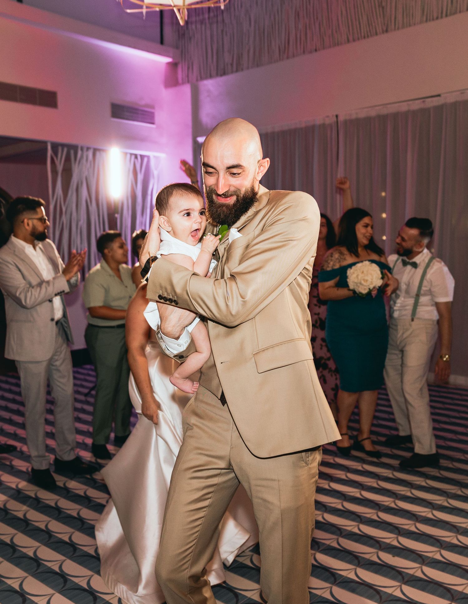 A person in a tan suit holds a baby while dancing at a wedding reception.