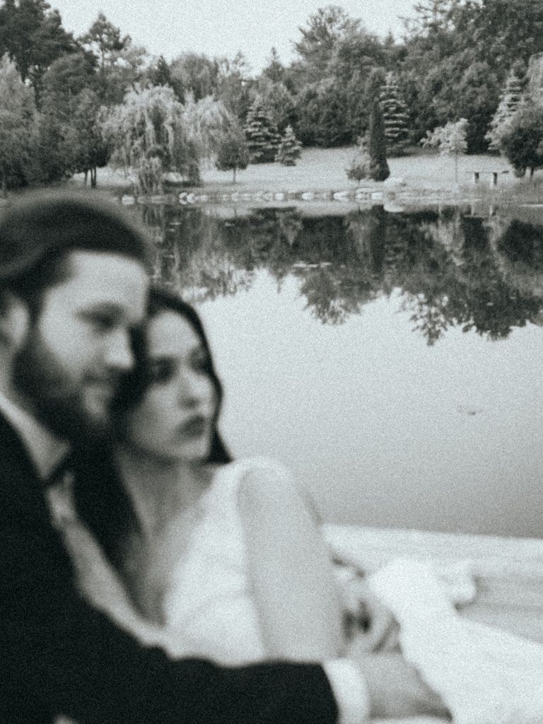 Black and white portrait of newlyweds by a serene pond with tree reflections.