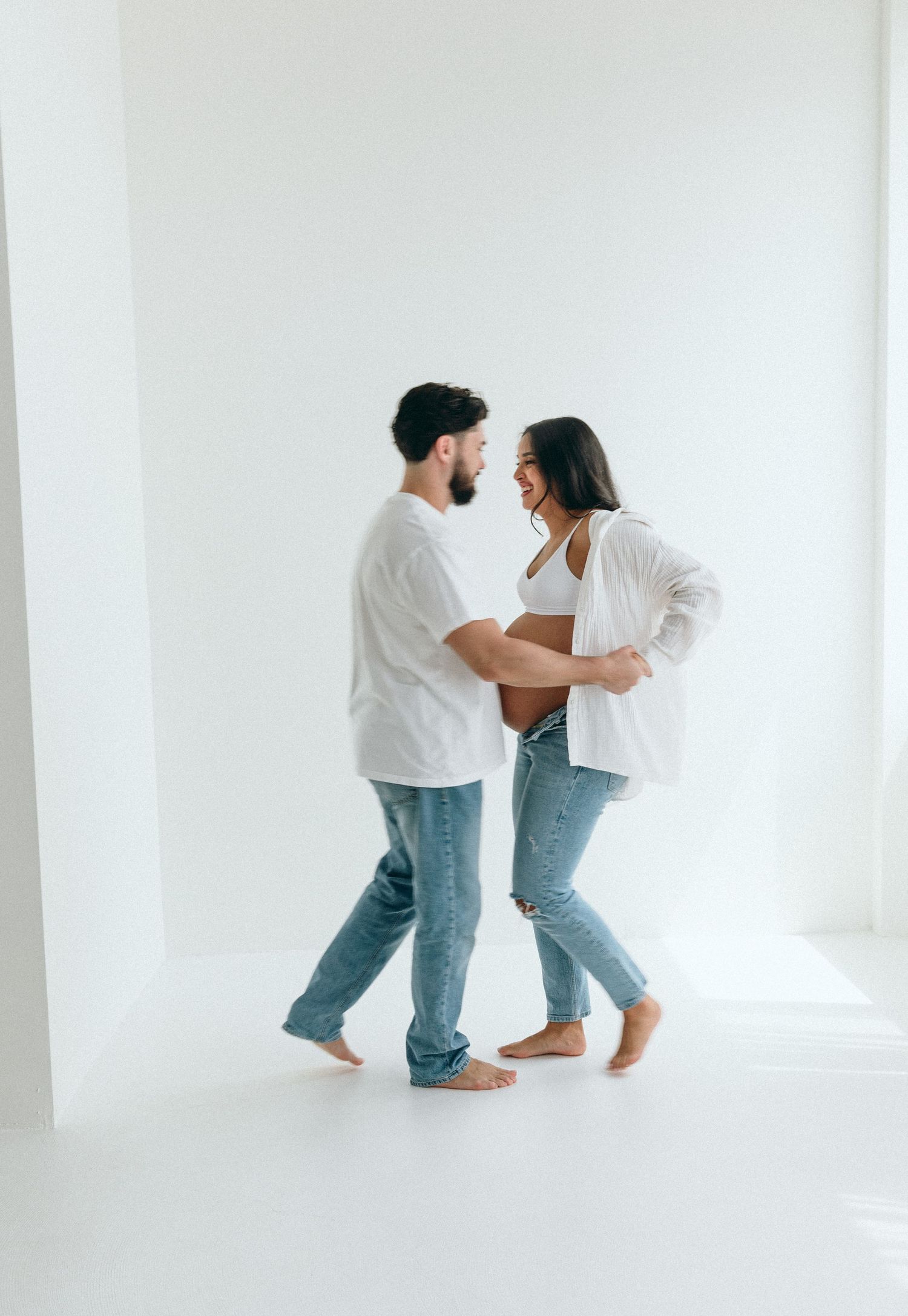 A couple in casual white tops and jeans dances playfully in an all-white room.
