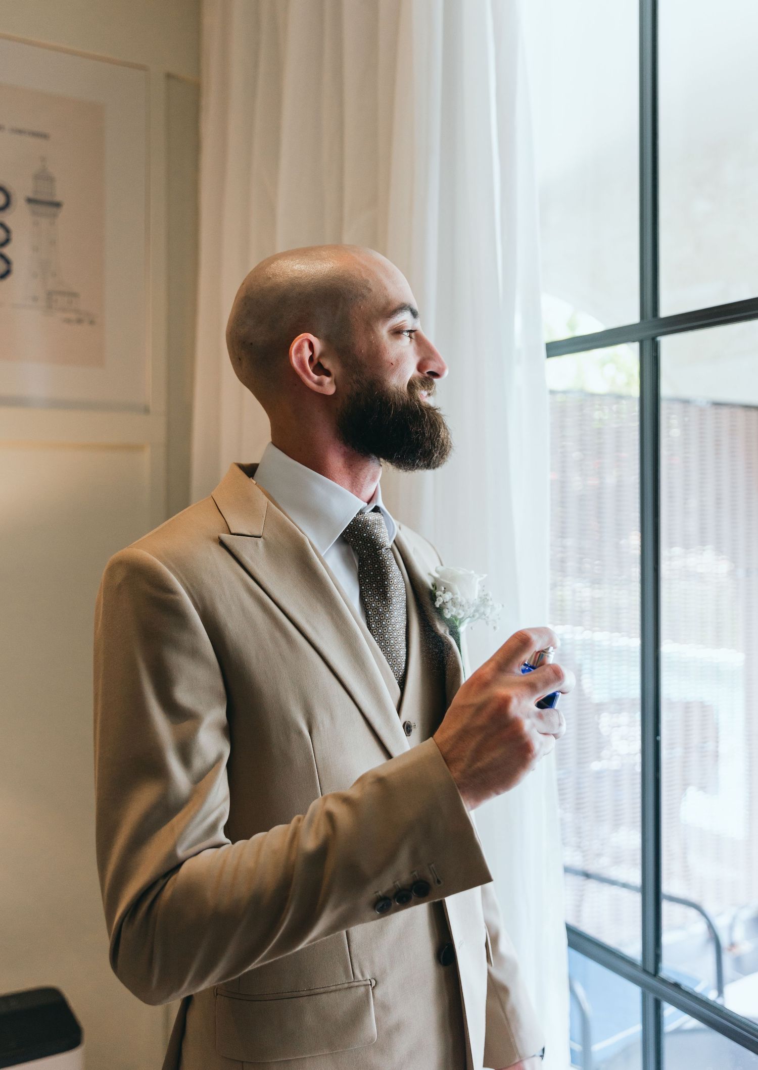 Person in beige suit jacket stands by window while drinking and gesturing.