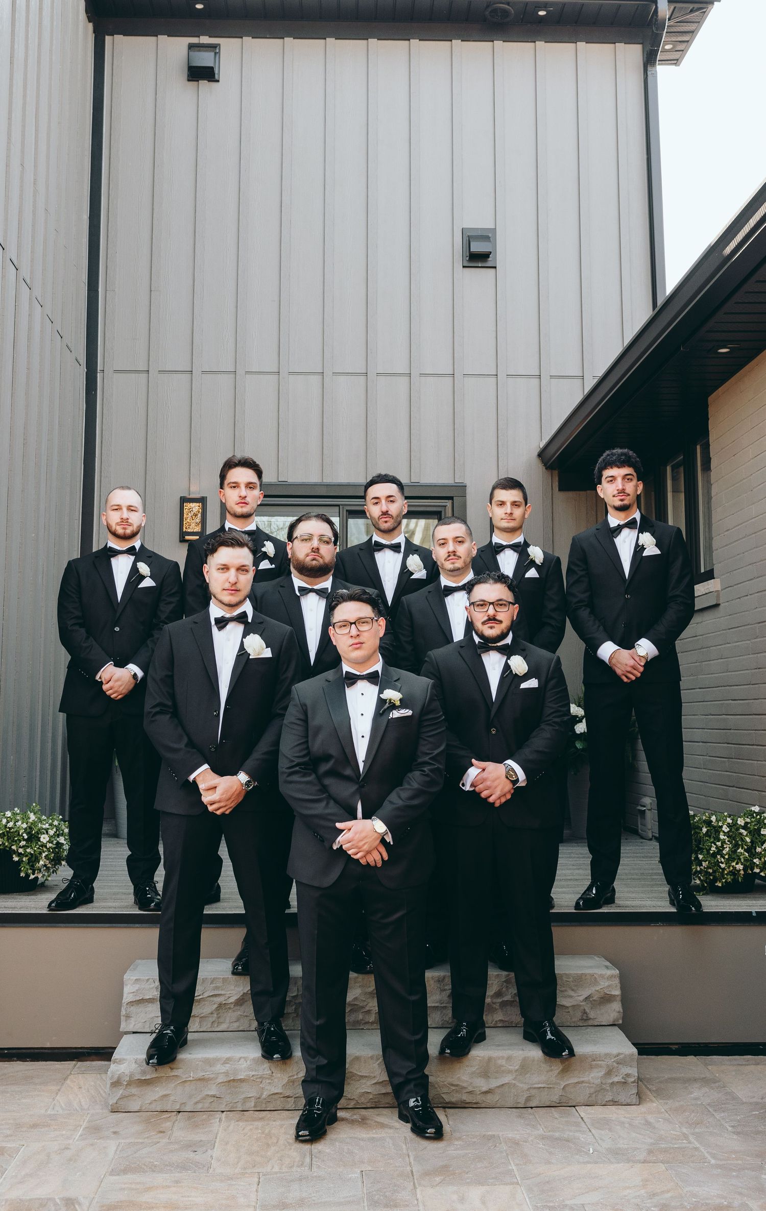 Wedding party of groomsmen in black tuxedos posing together outside a modern building.