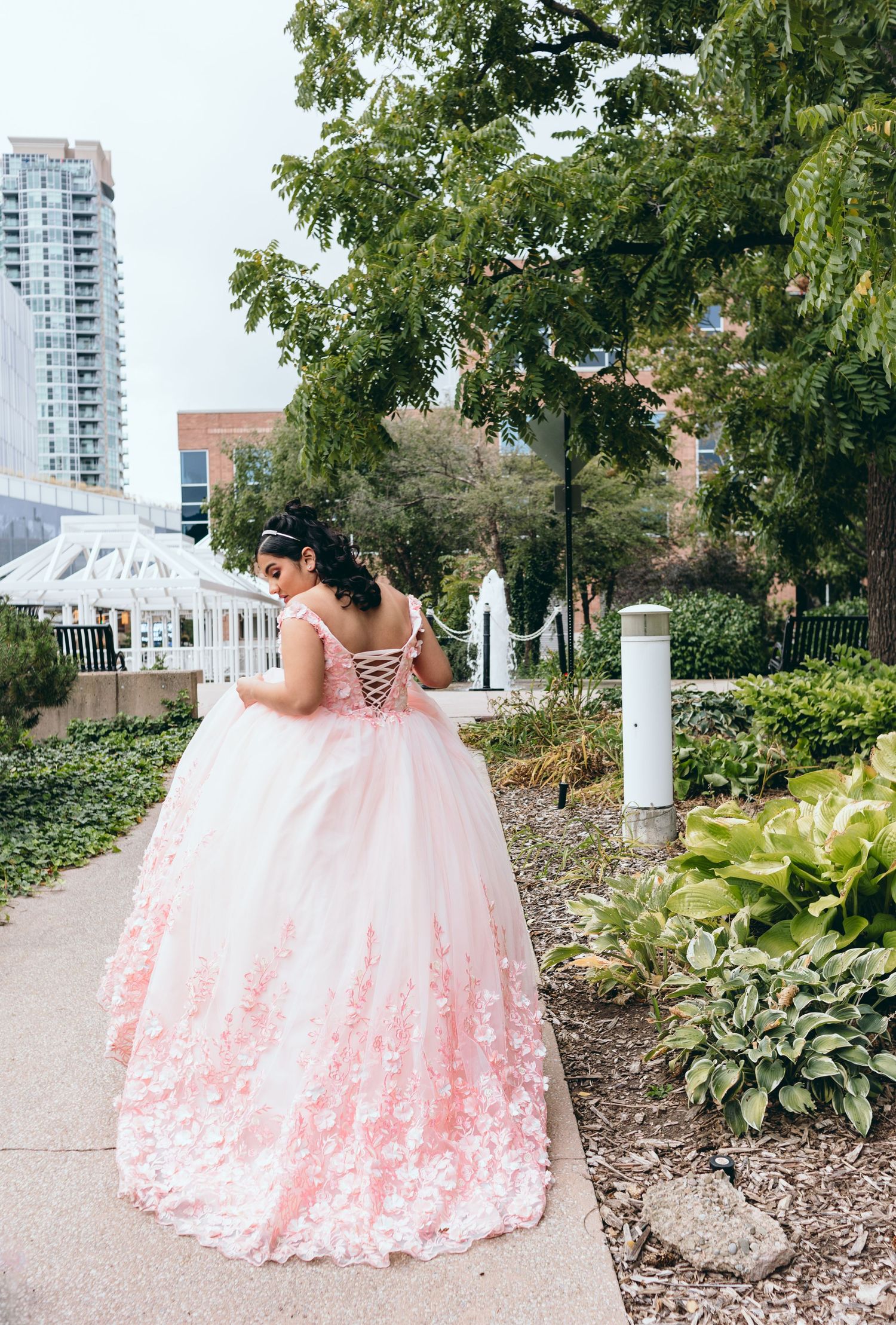 A pink ballgown with delicate lace details flows along a garden pathway with city buildings in the background.