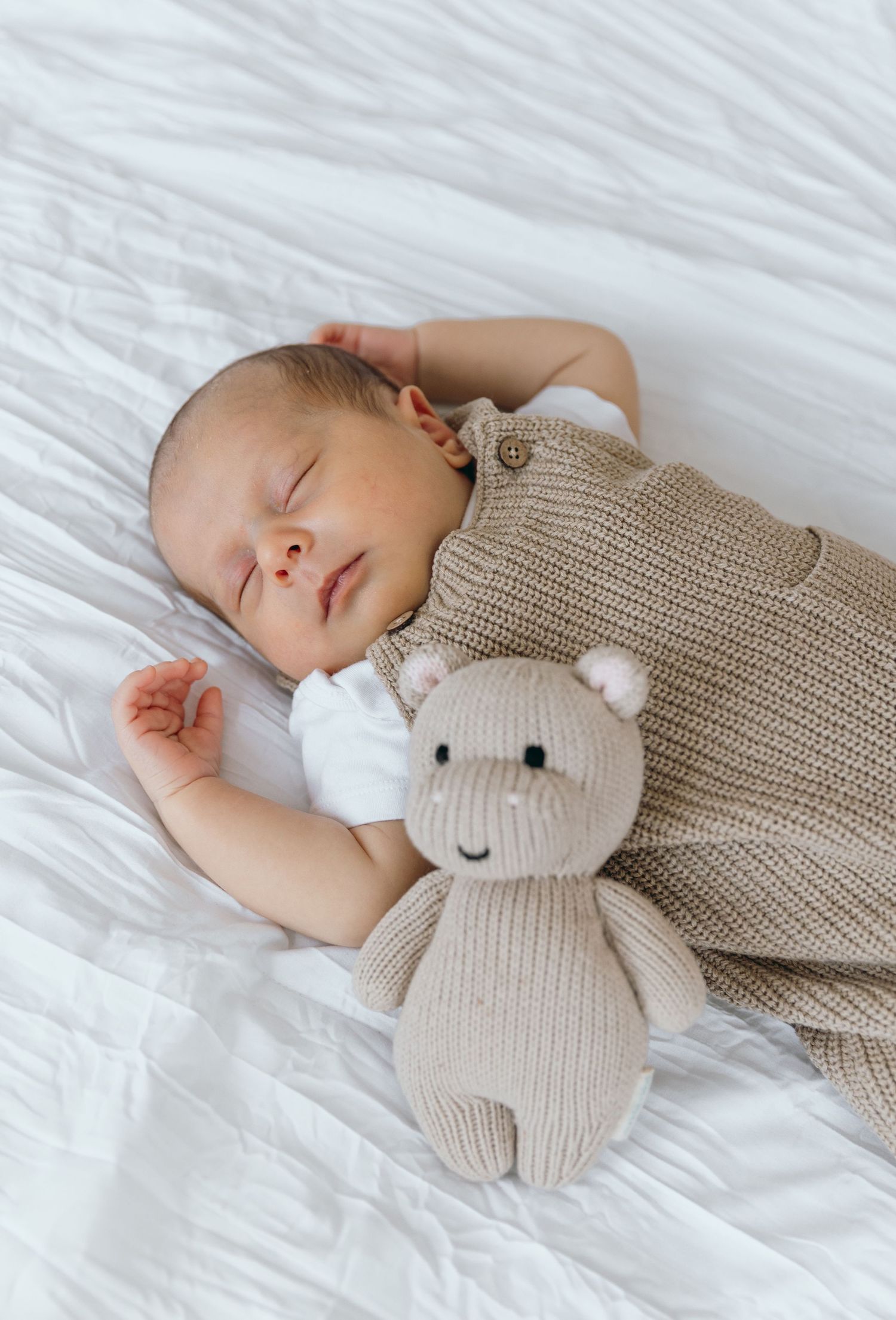 Baby napping on crisp white linens with beige knitted stuffed animal companion.