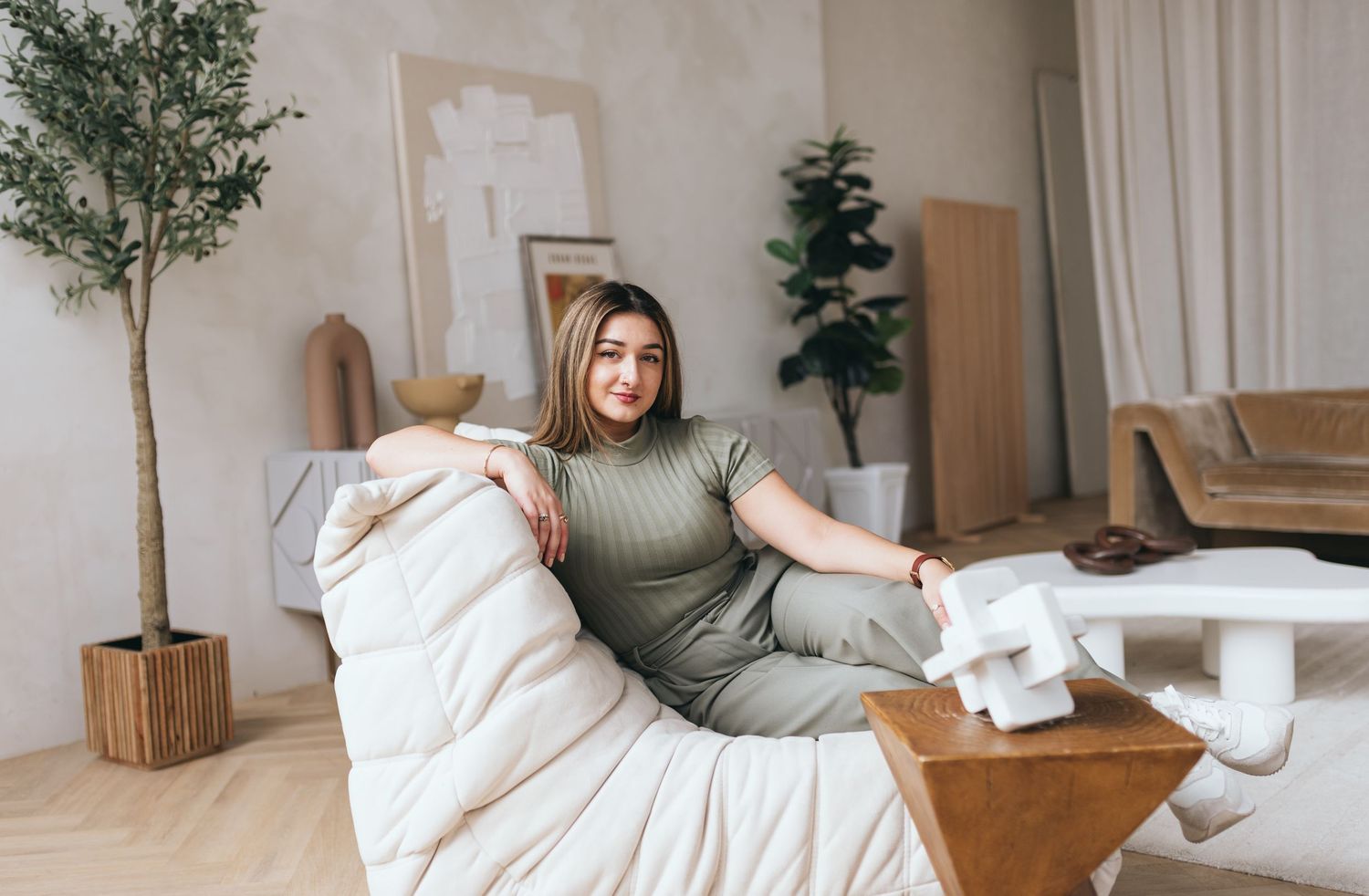 Woman in olive green sweater relaxing on cozy white bed with indoor plants in bright modern bedroom.