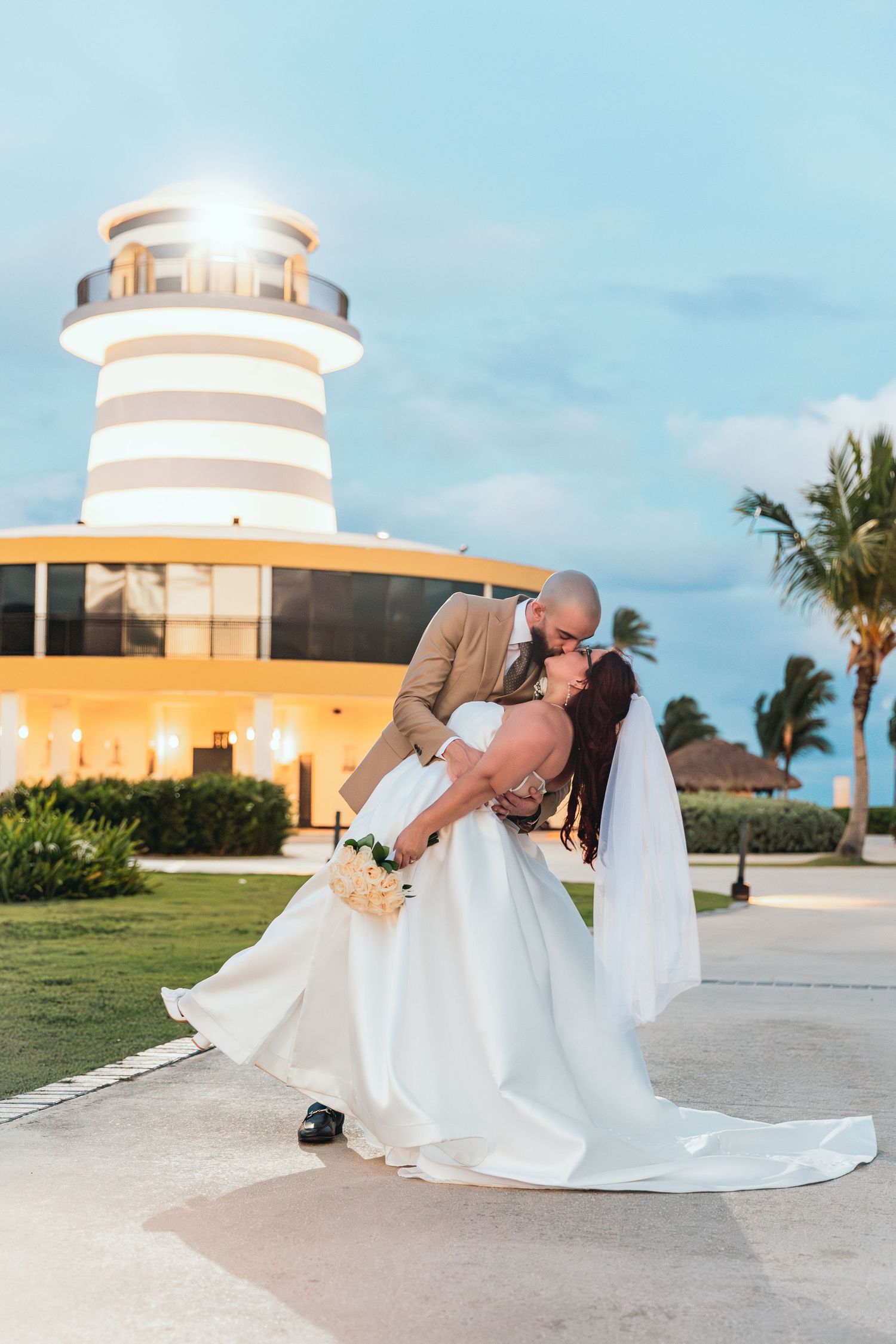 Newlyweds share a romantic kiss in front of a beige lighthouse building at sunset.