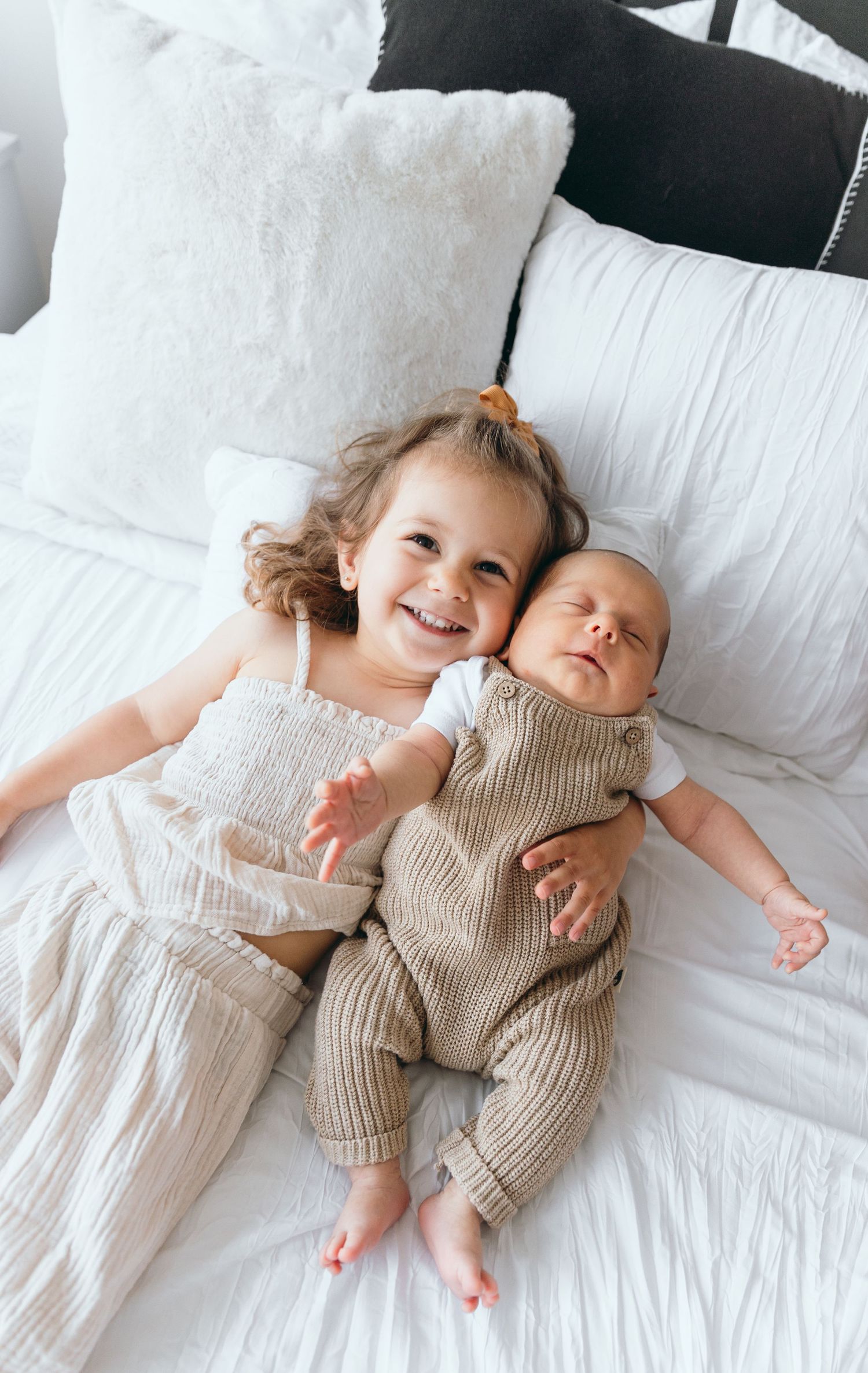 Two siblings relaxing together on white pillows with the older one smiling at the camera.