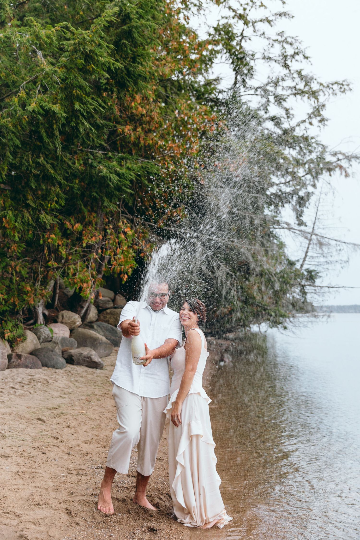 Playful couple in white outfits creating water splashes by the lake.