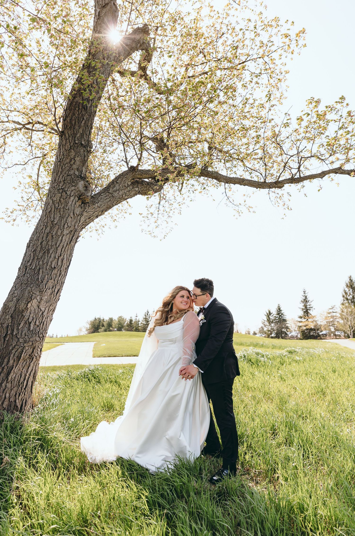 Wedding couple shares intimate moment under blooming tree in sunlit meadow.
