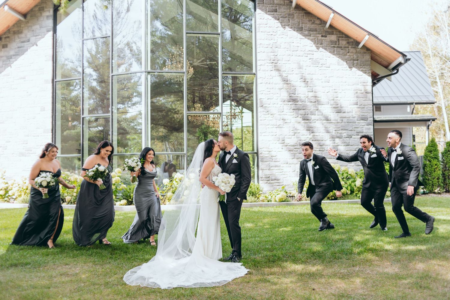 Wedding party jumps playfully behind newlyweds in front of glass chapel building.