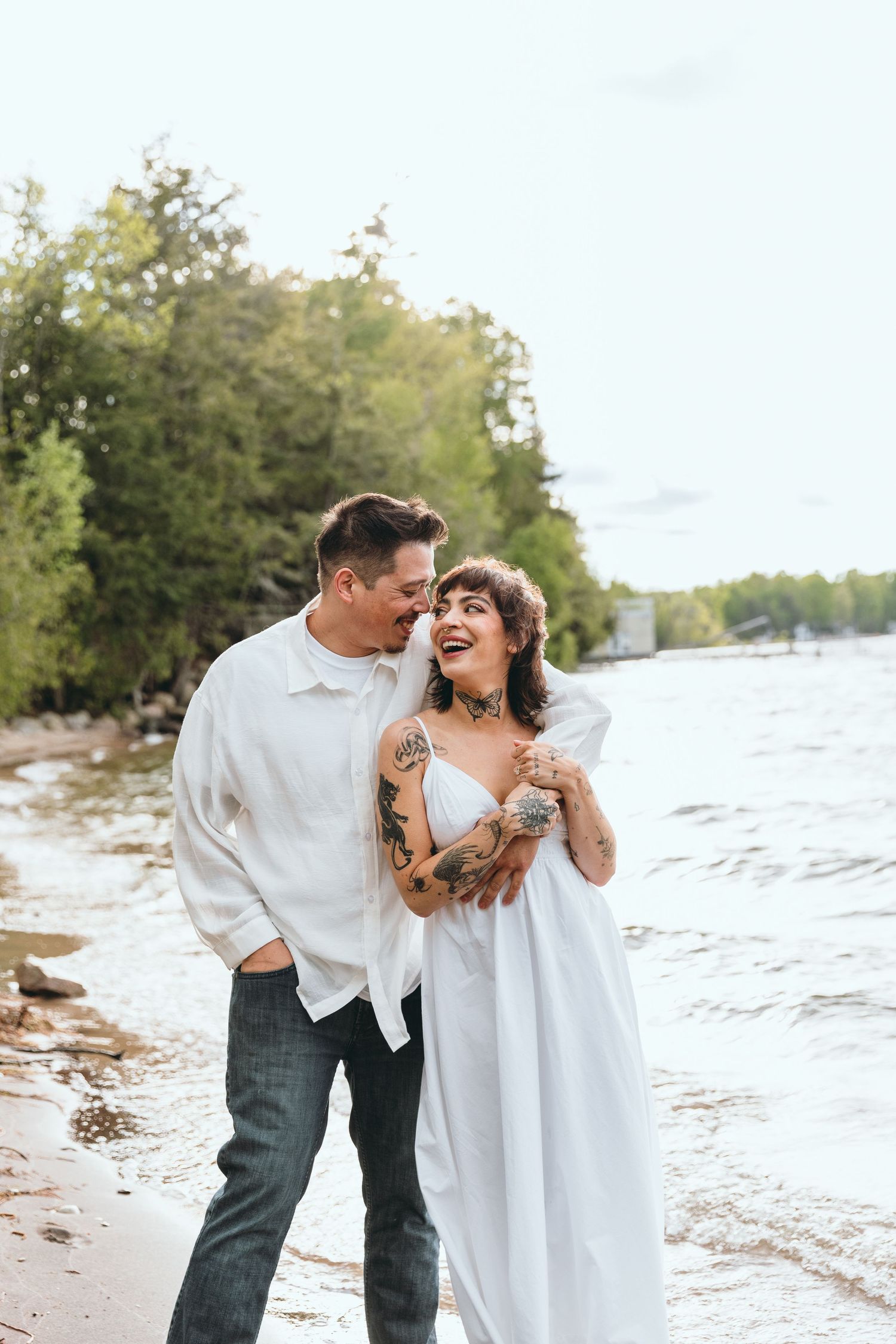 Joyful couple laughing together on beach shoreline in casual white outfits.