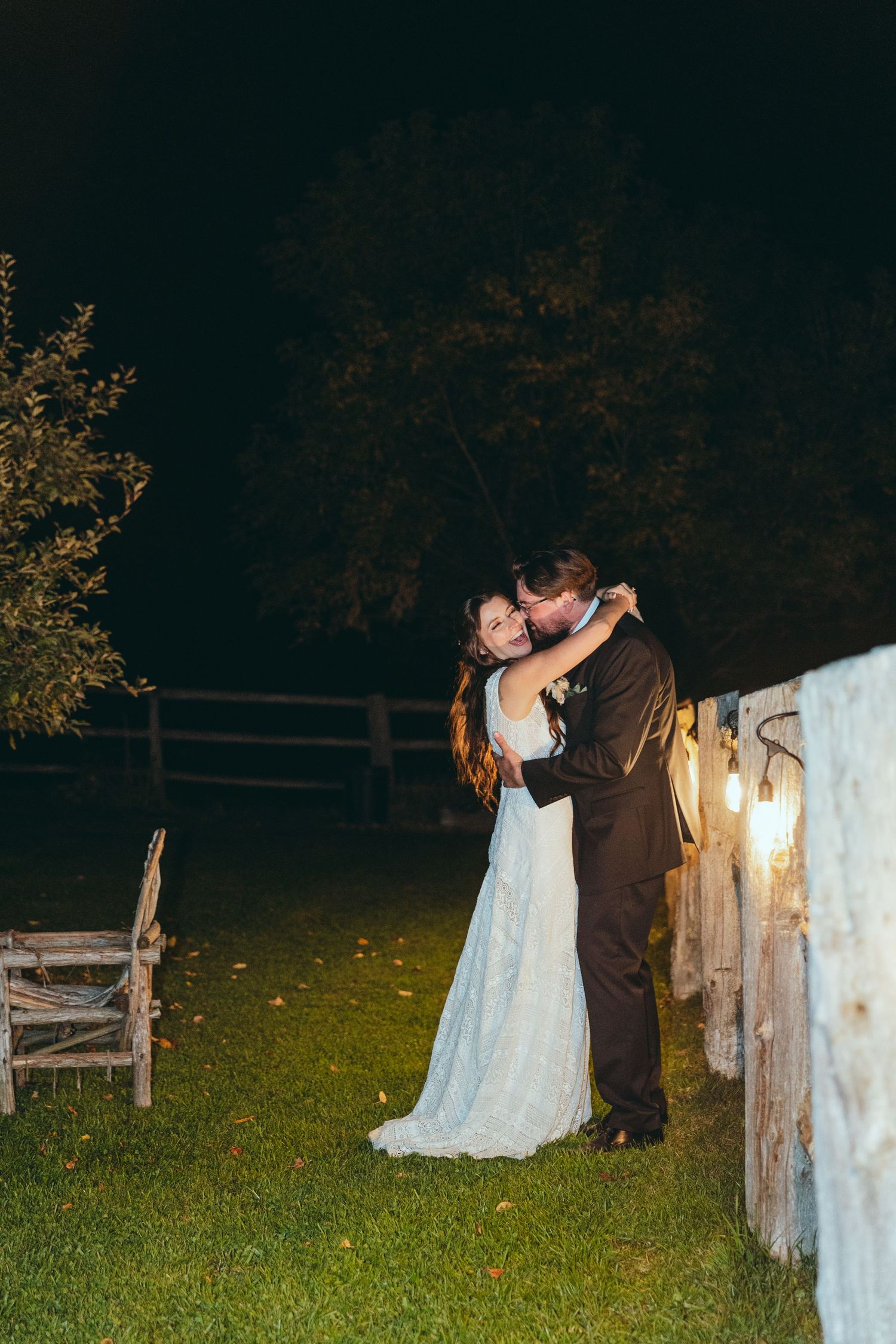 Nighttime wedding photos with couple embracing near rustic wooden fence and hanging lights.