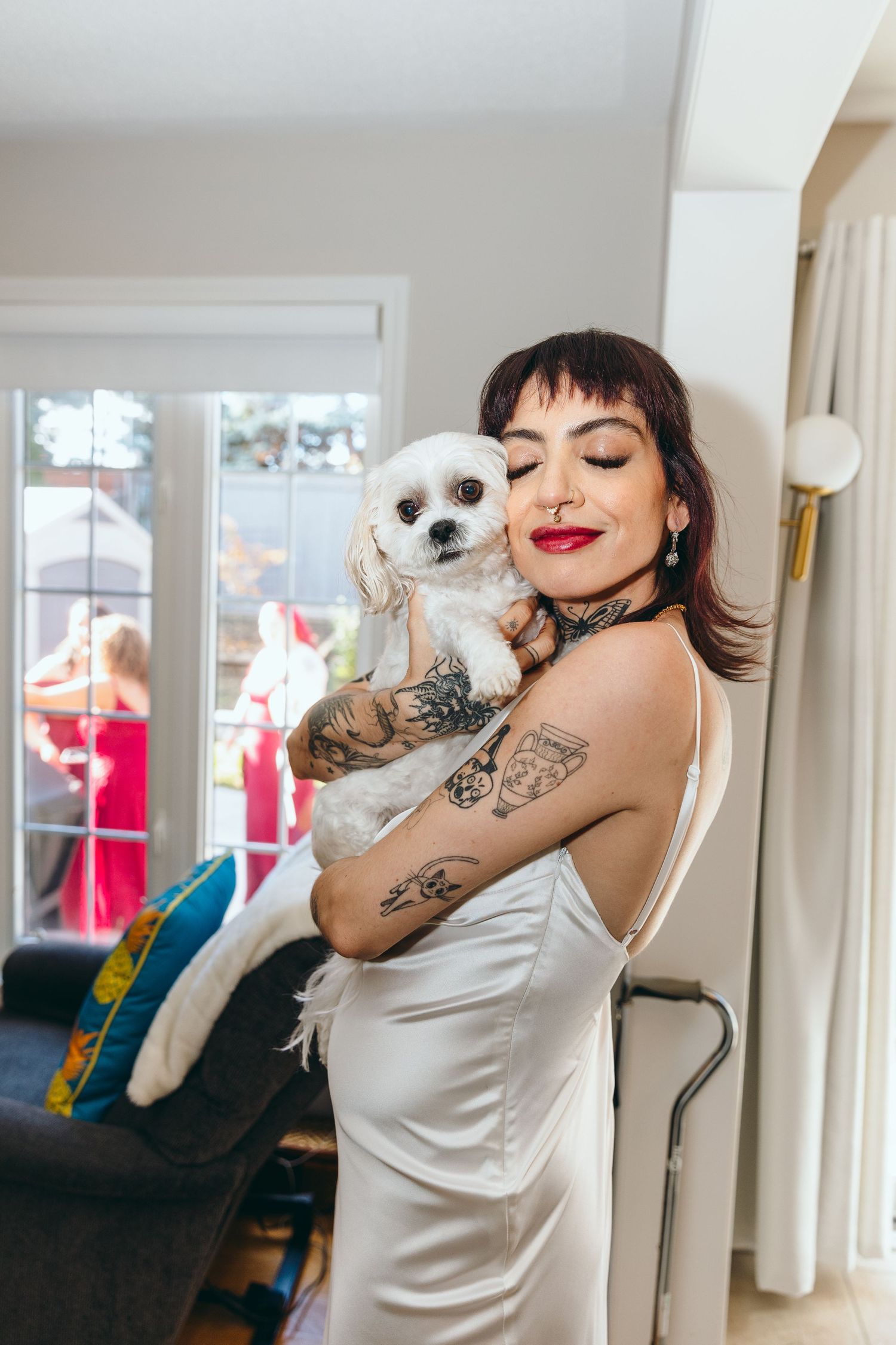 A person in a white silk dress holds and plays with a small white dog in a bright room.