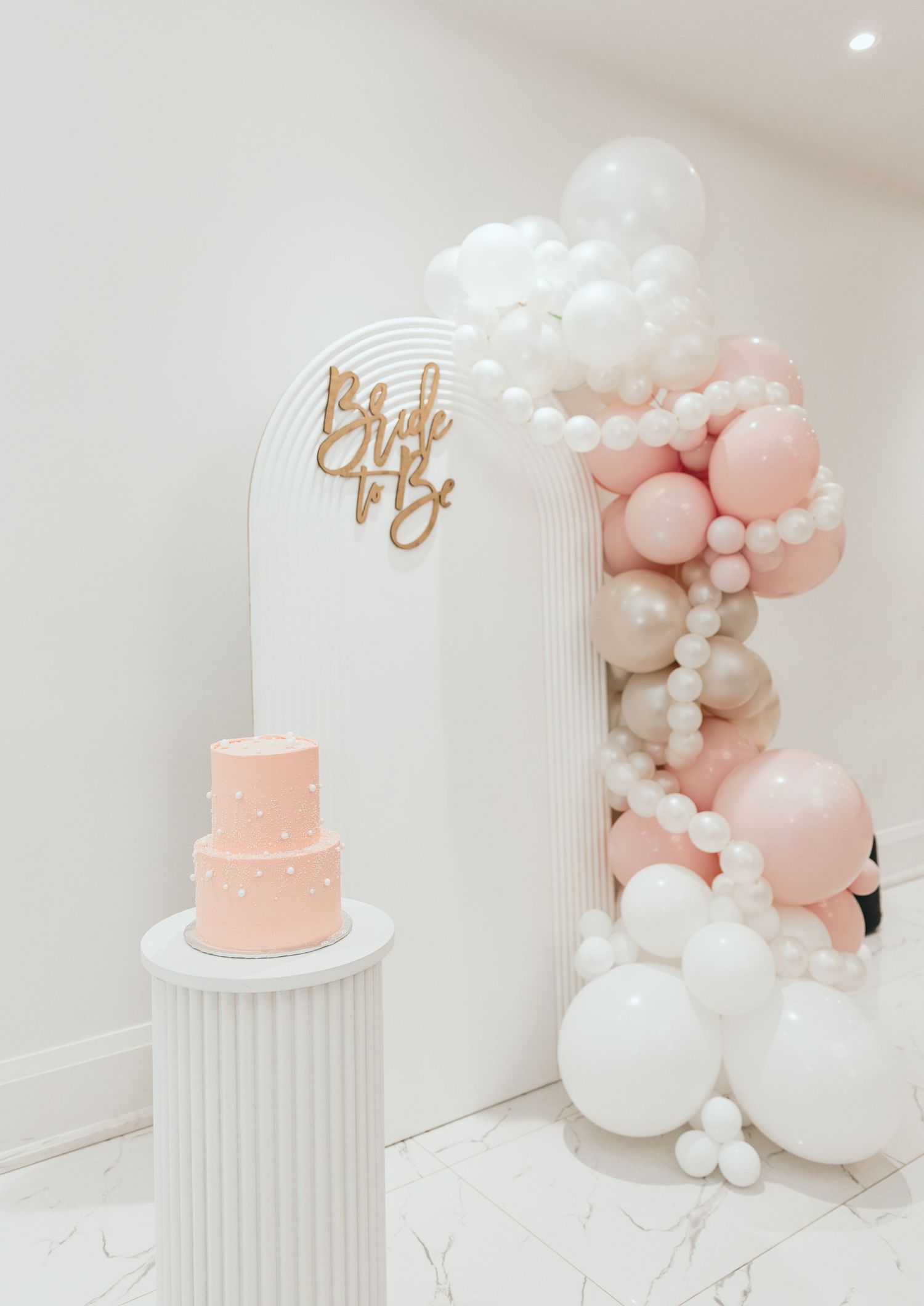 Pink and white balloon arch with elegant cake display on white pedestal.