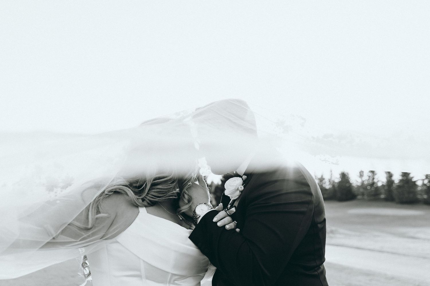Black and white photo of a couple embracing under a flowing veil outdoors.