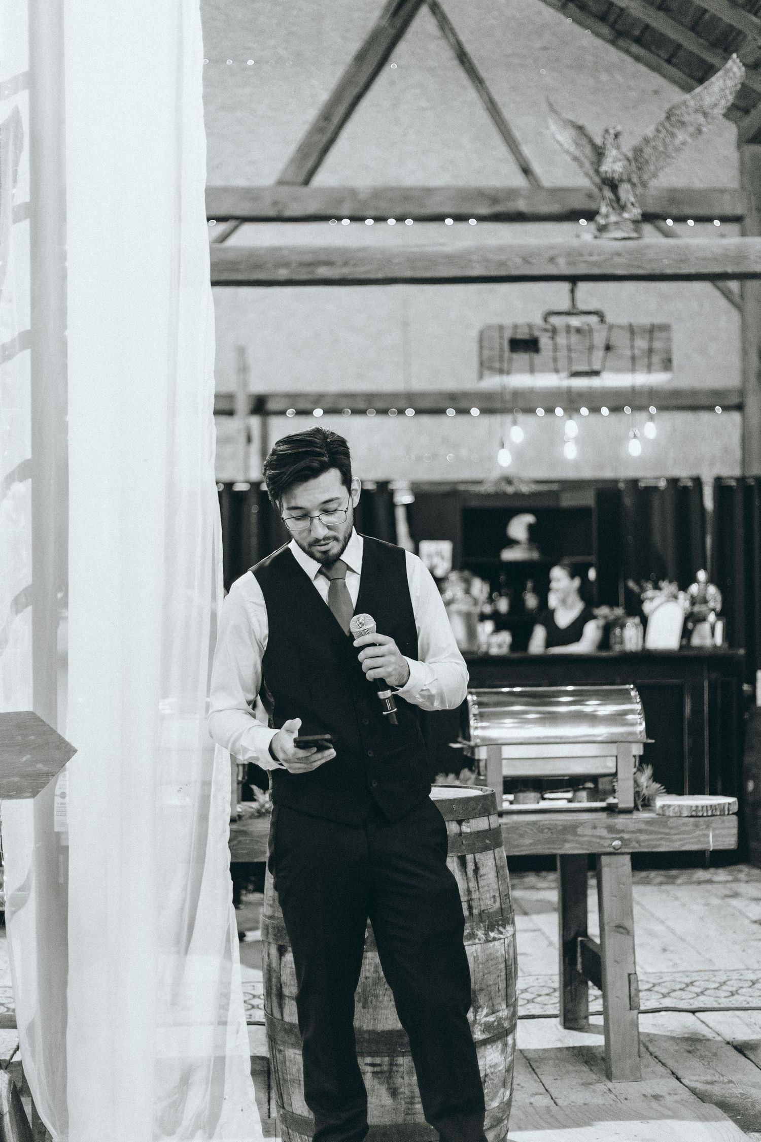 Wedding guests checking their phones during reception in rustic barn venue with white drapes and wooden beams.