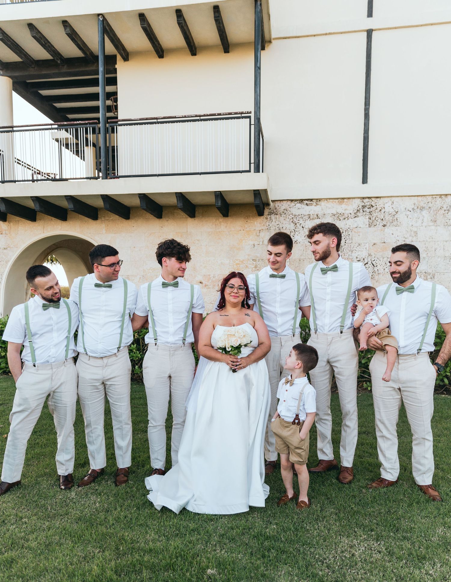 Wedding party in white attire poses playfully on grass in front of building.