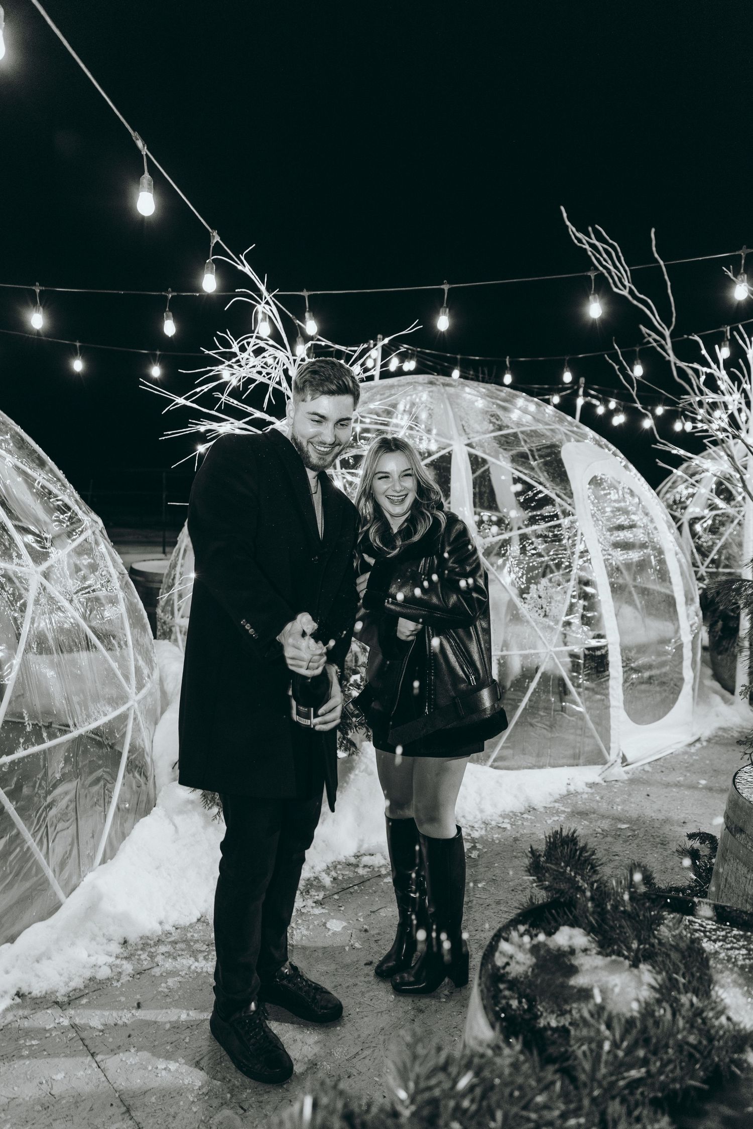 A couple poses in front of illuminated igloos during a snowy winter evening.