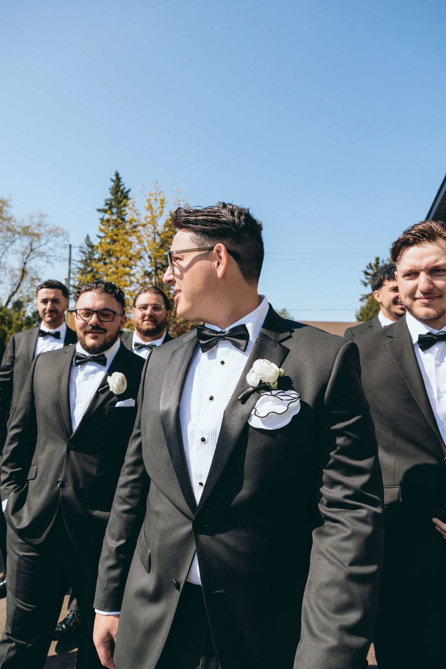 Groomsmen in black tuxedos and bow ties gather outdoors for wedding photos.