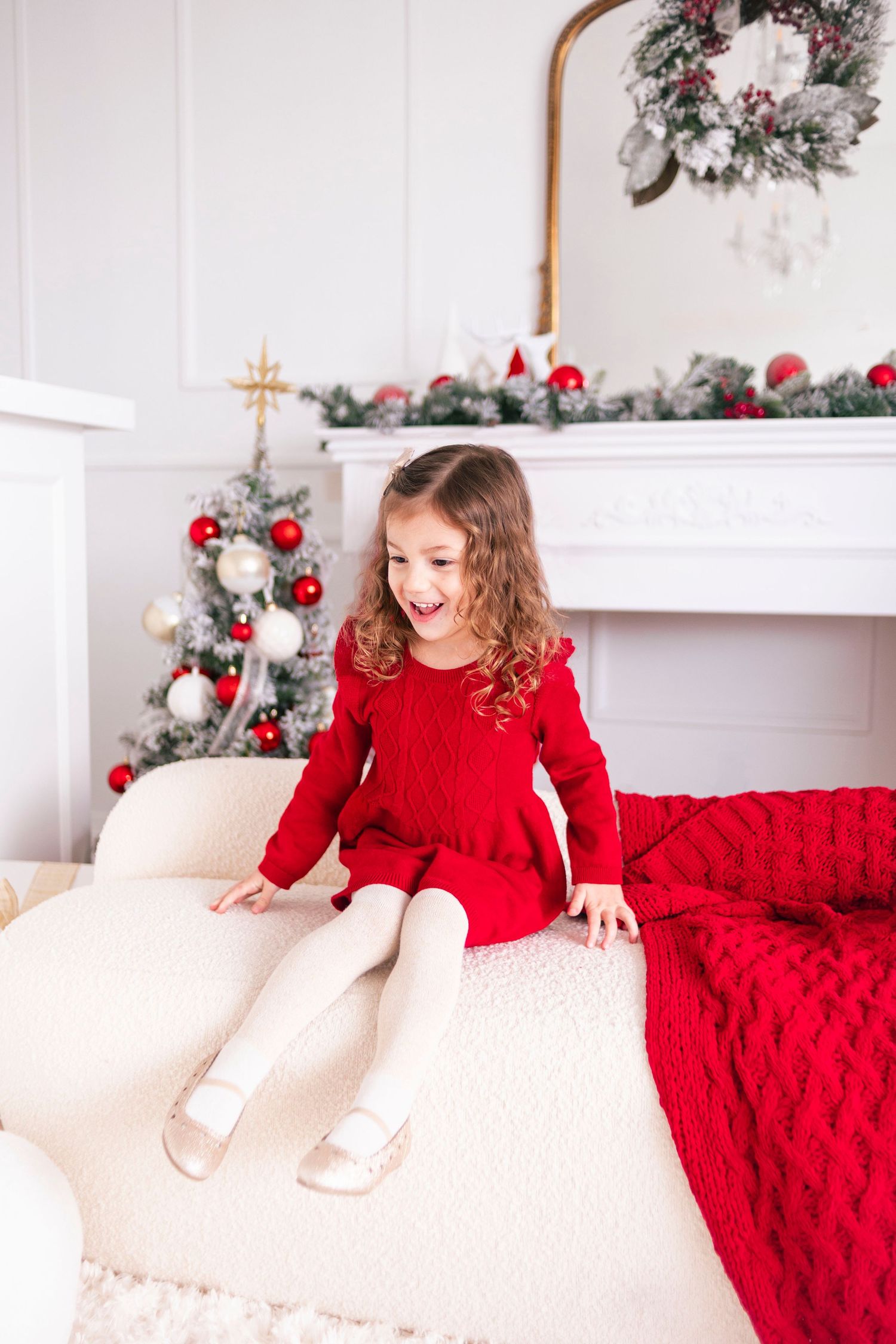 A joyful little girl in a red knit dress sits on a white sofa near a decorated Christmas tree and fireplace.