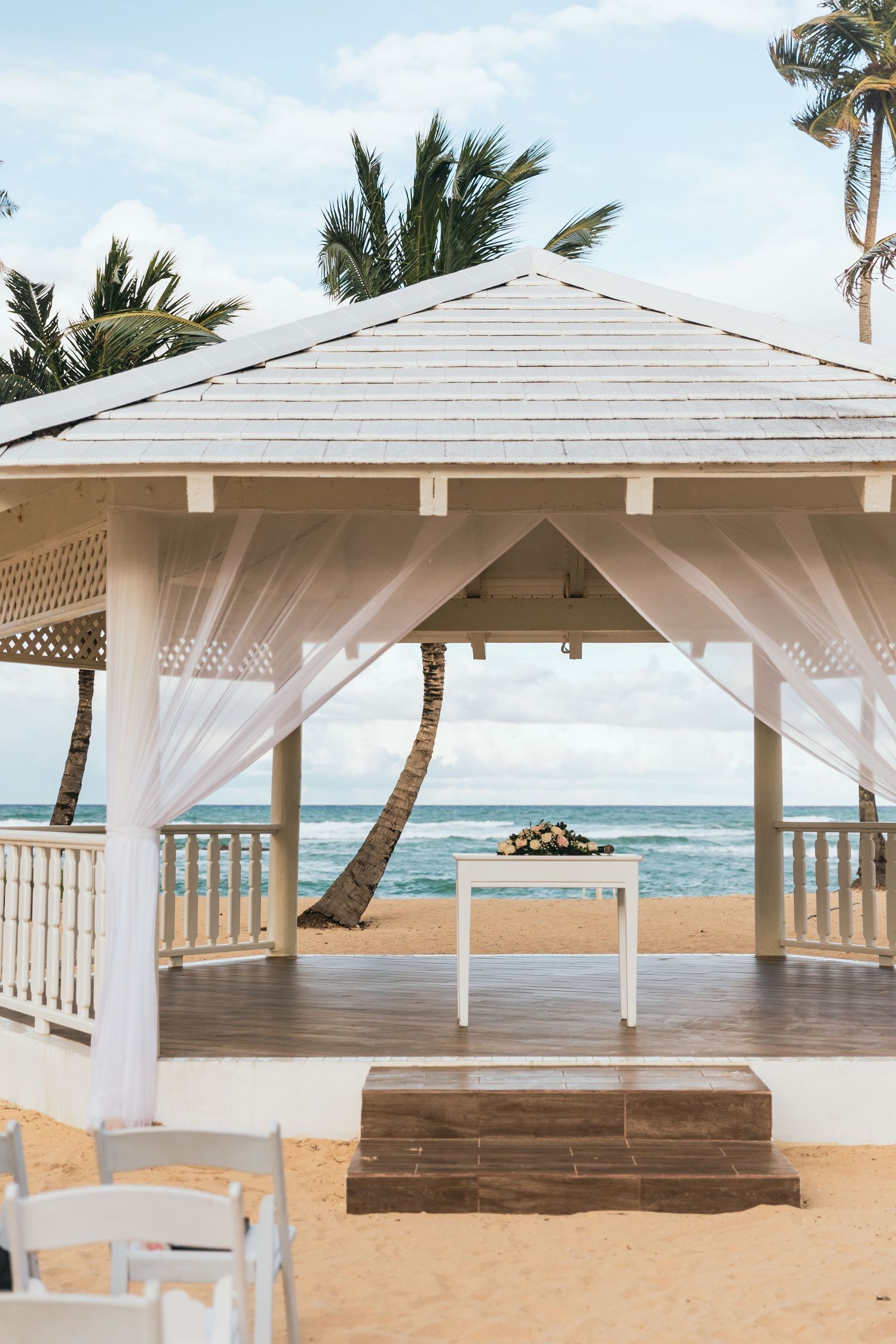 White beachside wedding pavilion with flowing curtains and wooden benches overlooking ocean.