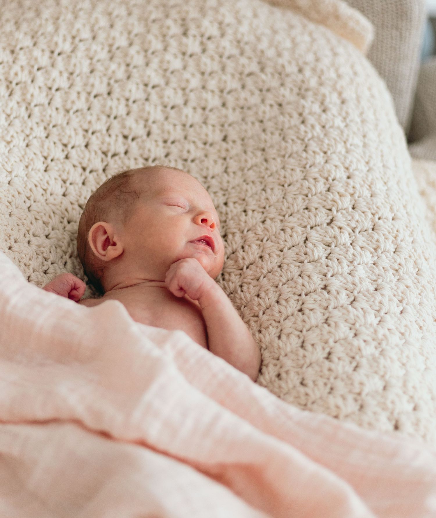 Newborn baby resting on textured cream-colored blanket in soft natural light.