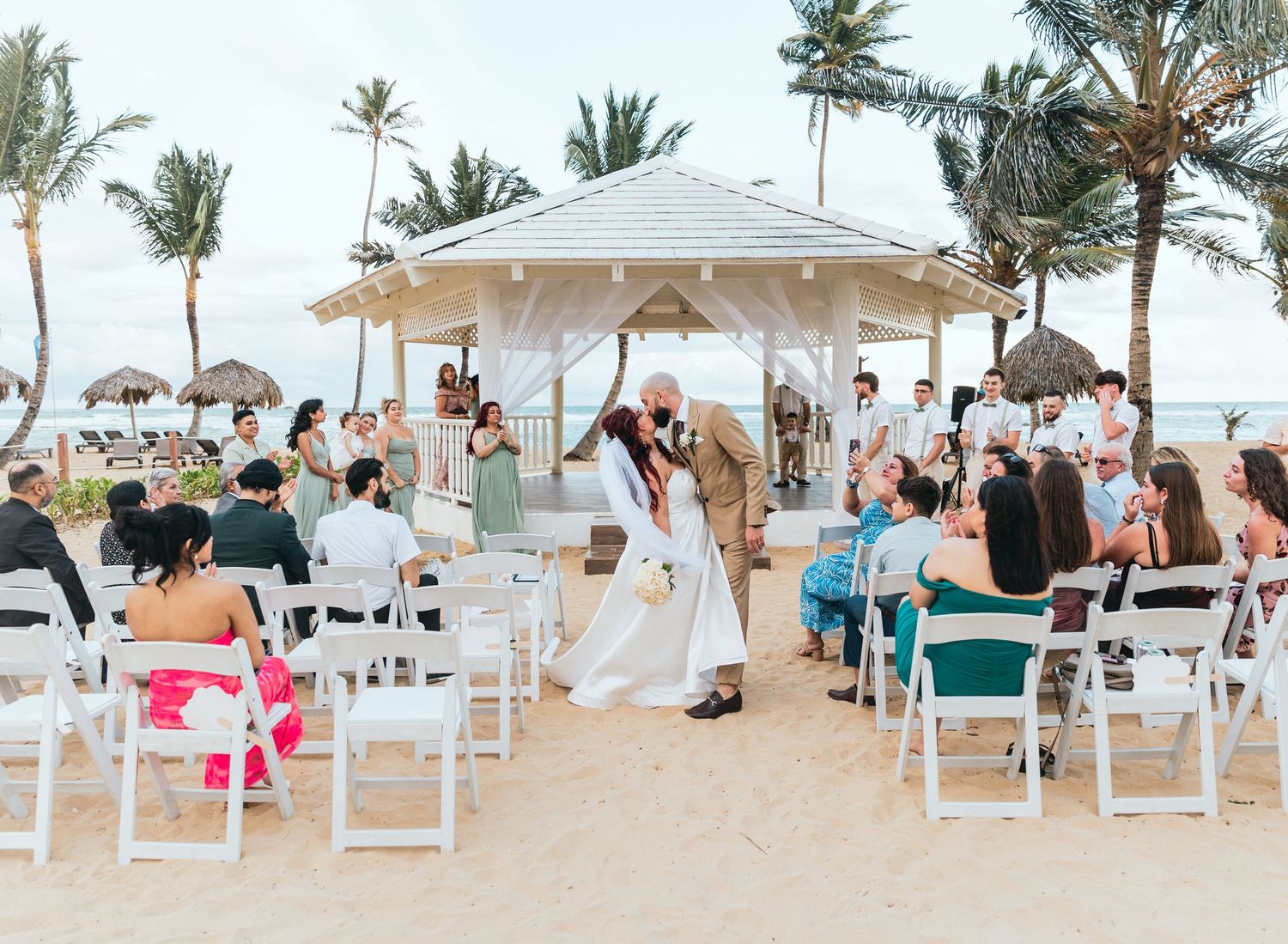 Wedding guests seated in white chairs watch a ceremony under a beachside gazebo with palm trees.