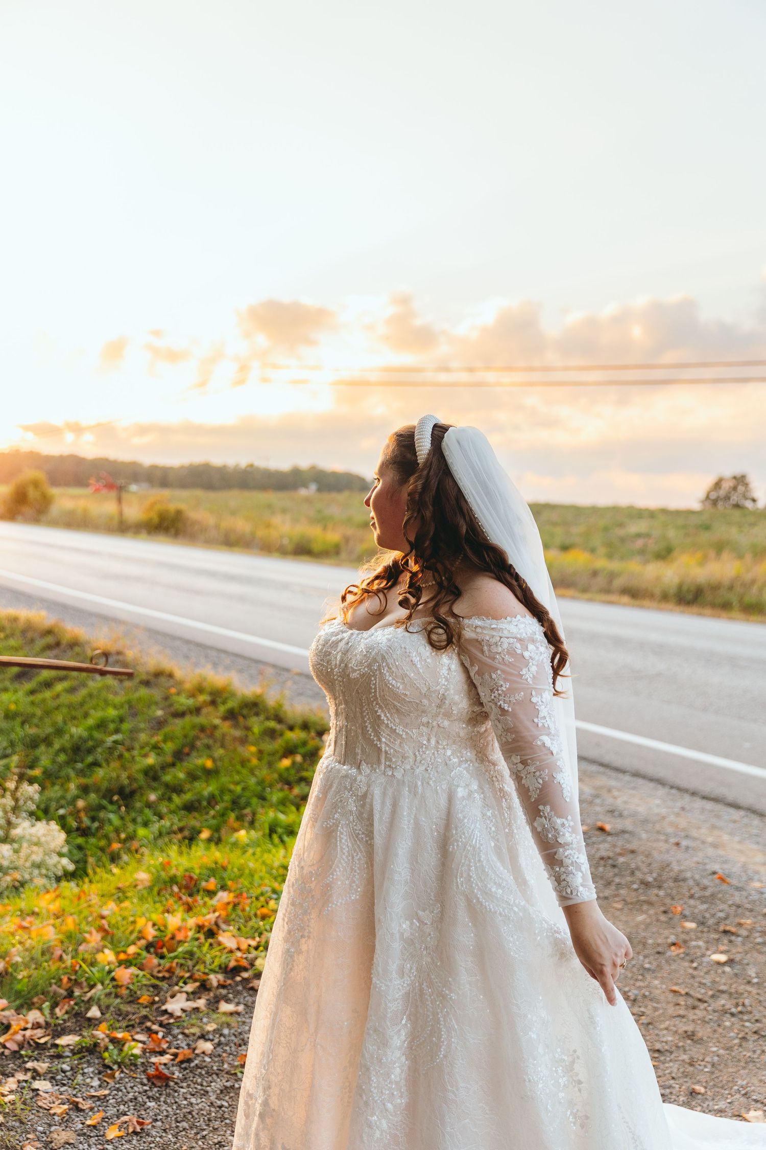 Bride in lace wedding dress walks along rural road at sunset.