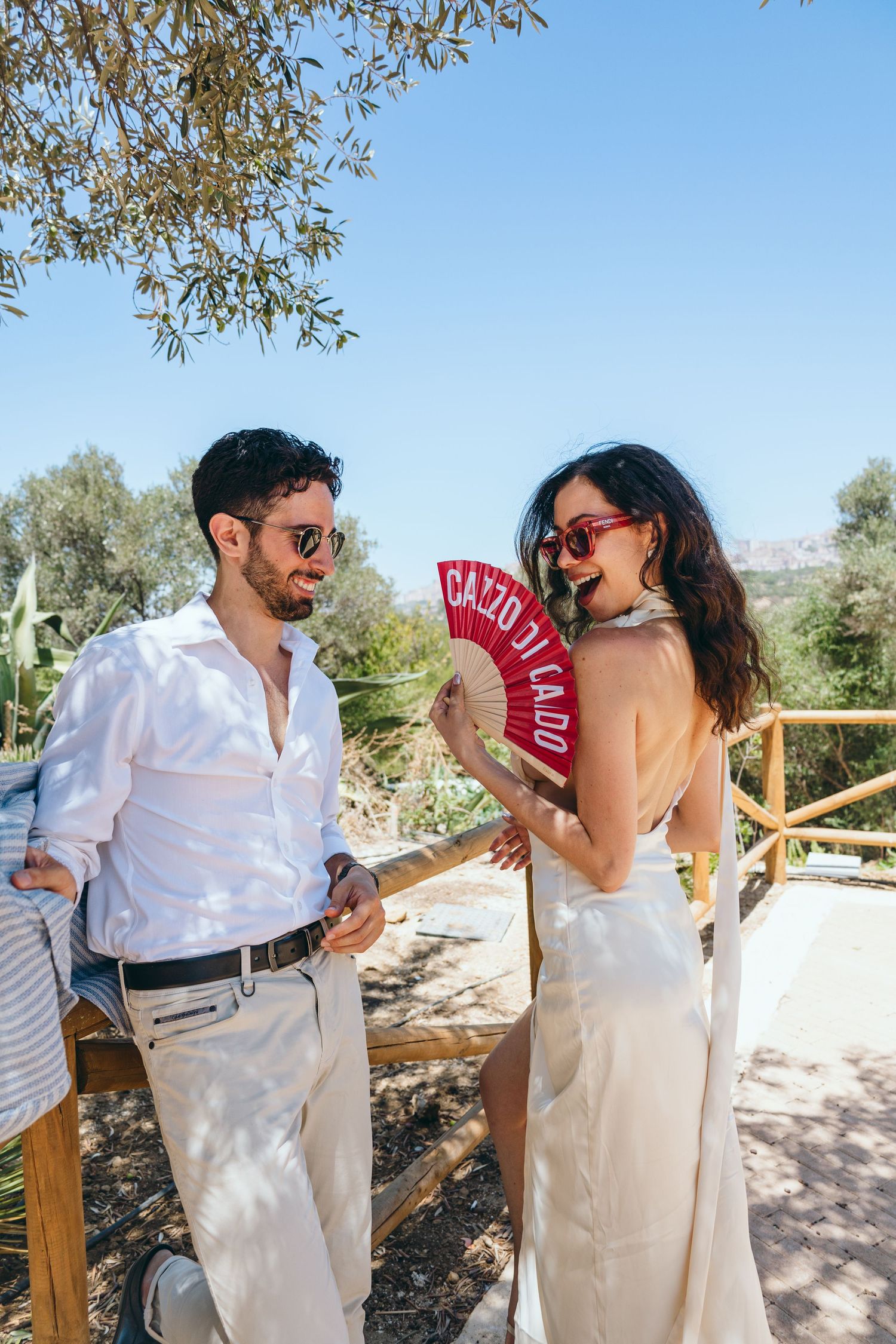Couple in summer white attire share playful moments with red fan in garden setting.