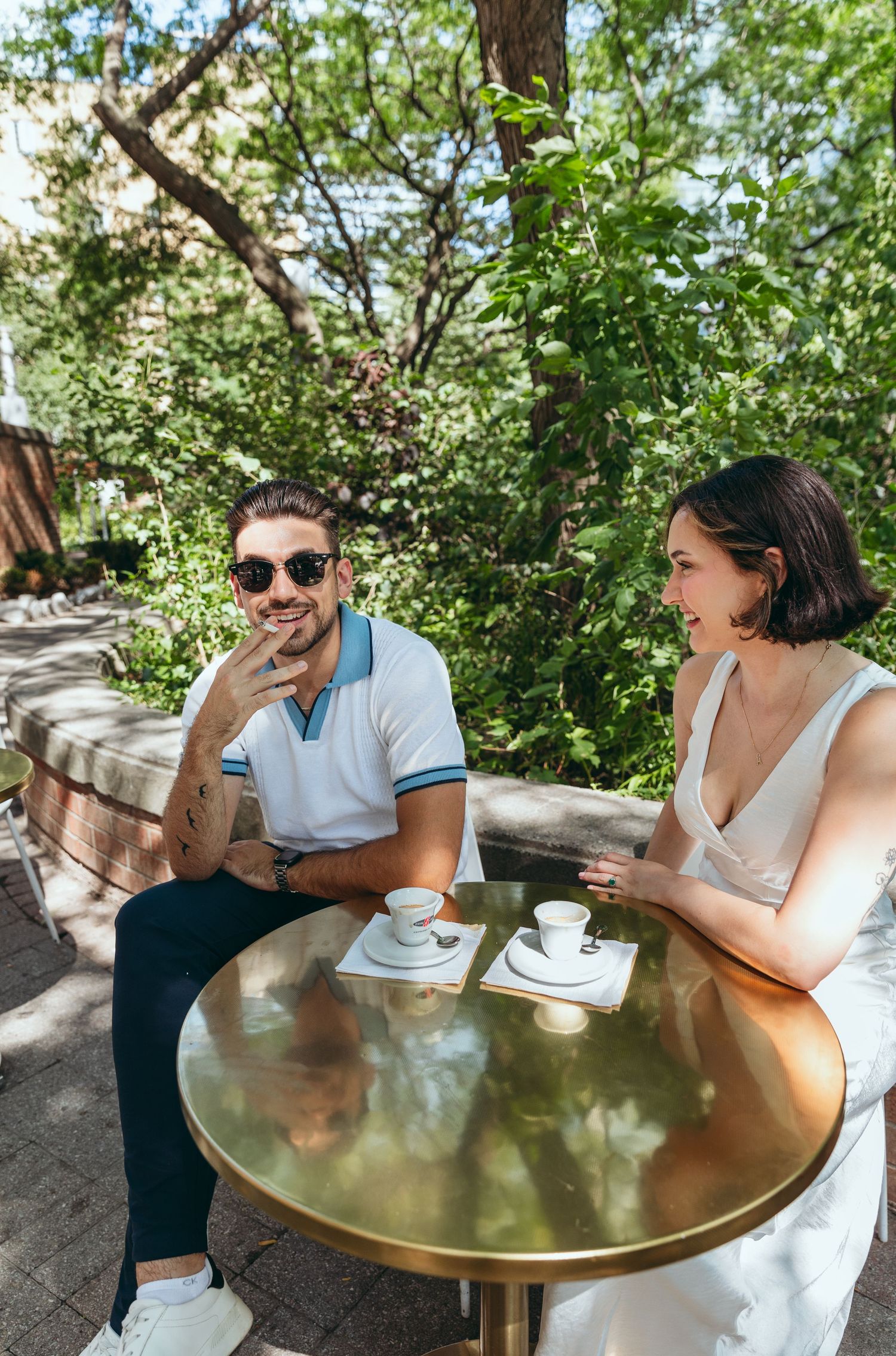 Two people share a lively conversation while seated at a round brass table outdoors.