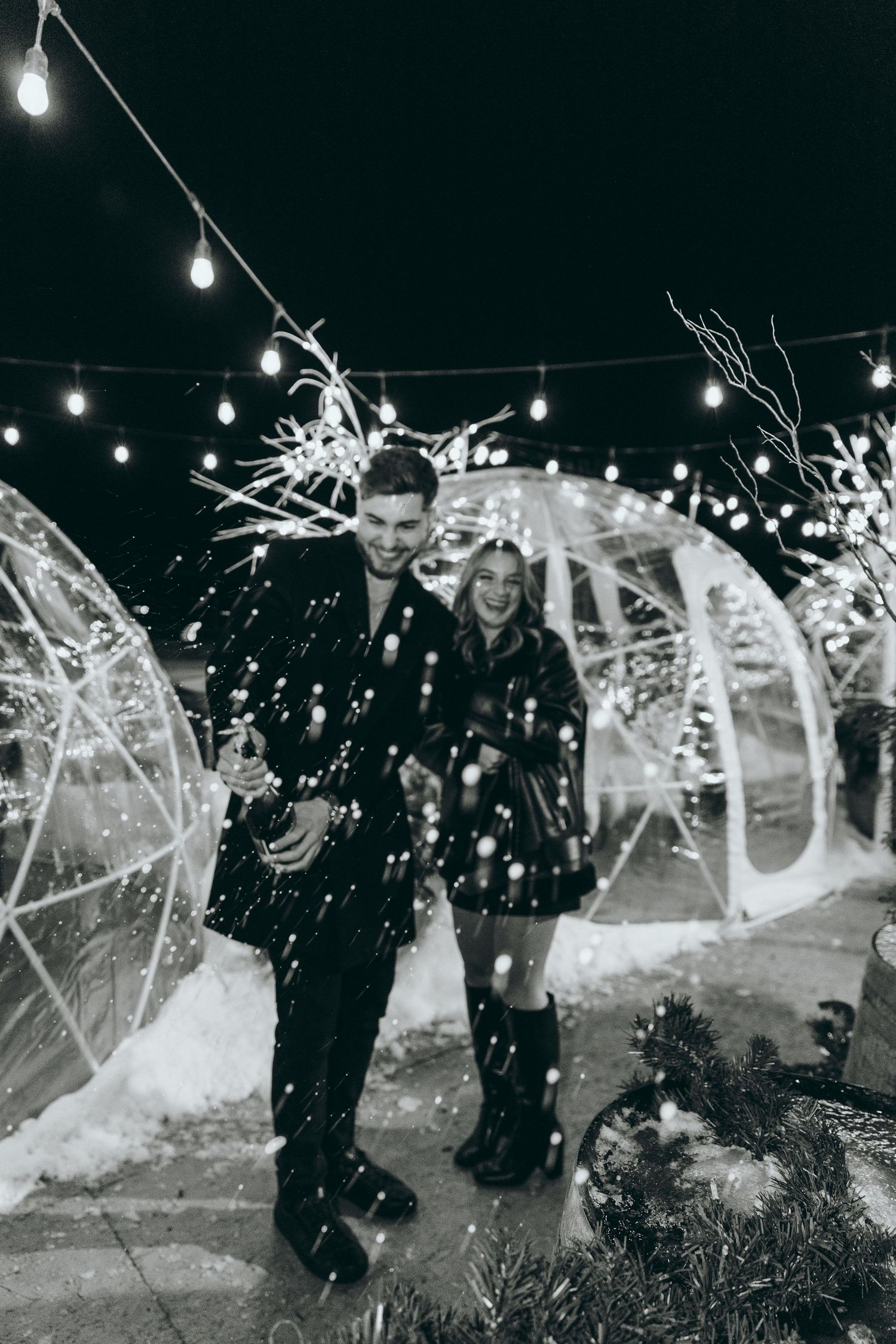 A couple stands in falling snow near illuminated dome structures at night.
