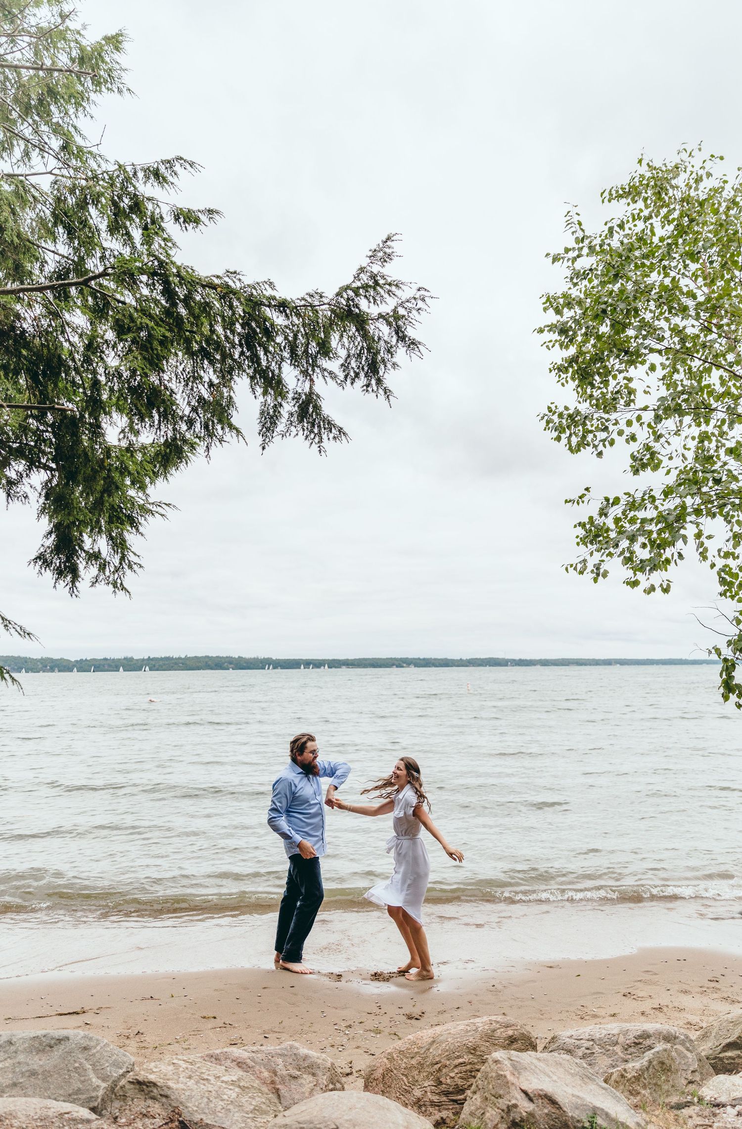 A couple dances playfully on a beach with evergreen trees and a lake view.