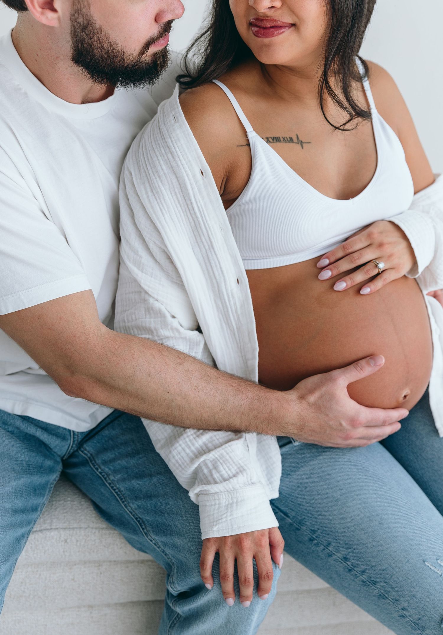 A tender maternity moment showing hands cradling a pregnant belly while wearing casual white and denim clothing.