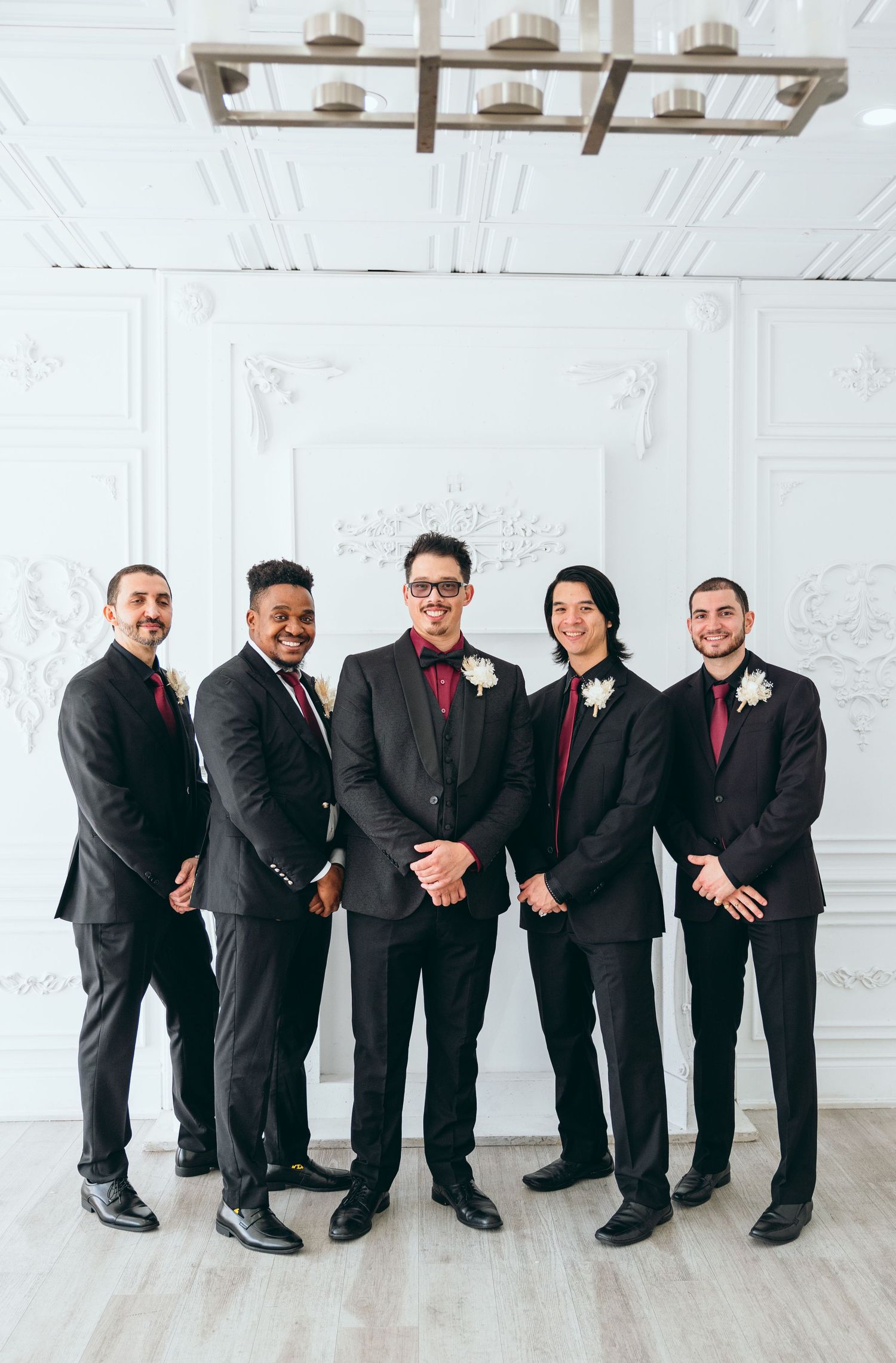 Groomsmen in black suits with red accents posing in front of a white decorative wall.