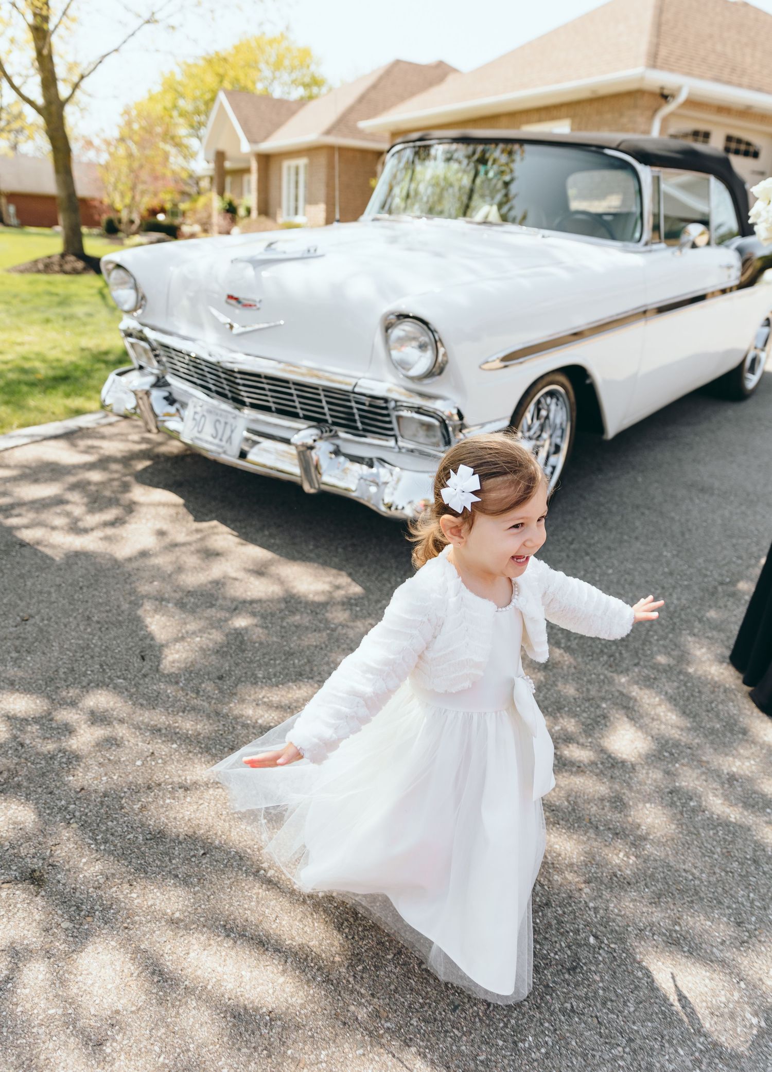 A young flower girl in white dress twirls in front of a vintage Chevrolet.