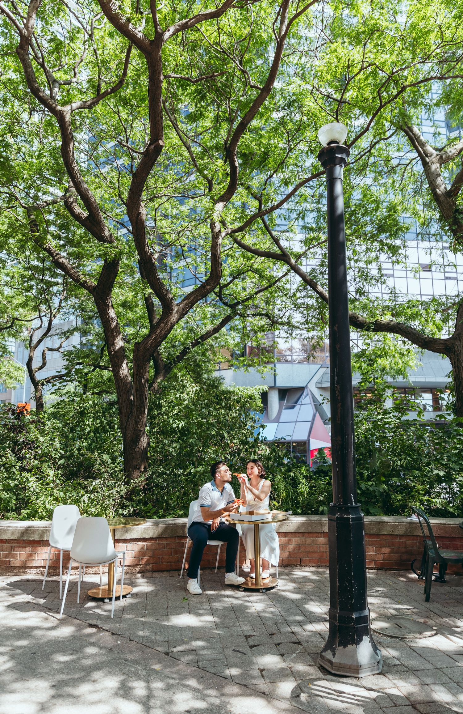 Wide shot of a tree-lined outdoor plaza with a couple sitting at a distant table.