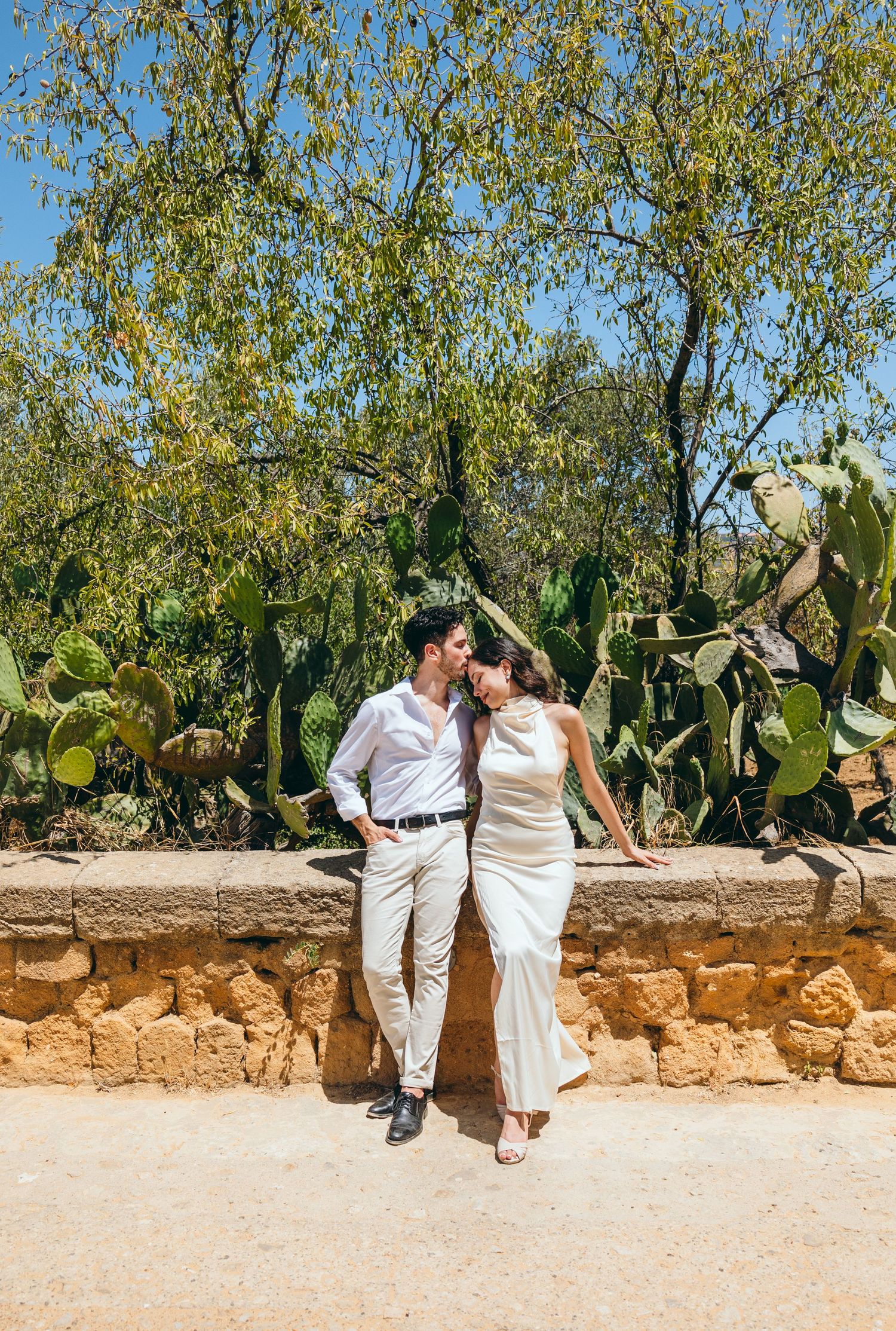 Romantic couple in matching white outfits pose against stone wall with desert garden backdrop.