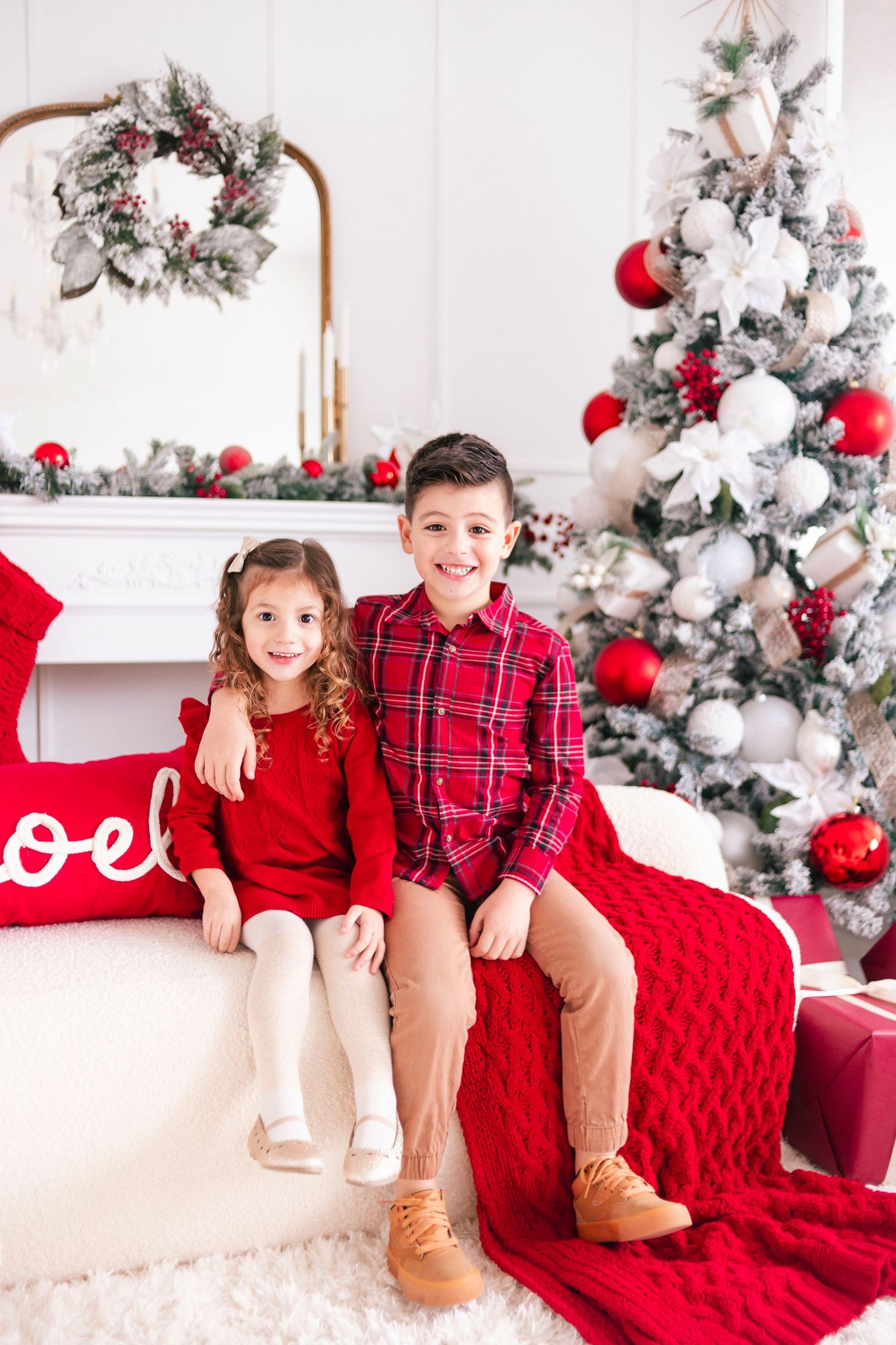 Two children in red outfits sitting on a sofa with Christmas tree and festive decorations in background.